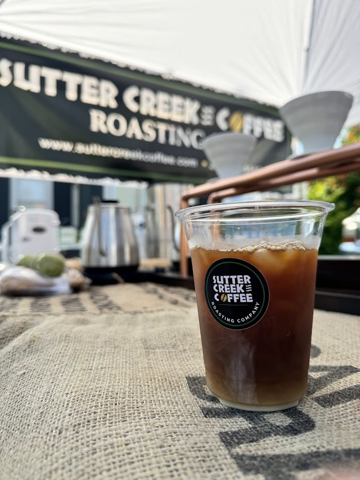 Plastic cup filled with iced coffee on a burlap tablecloth, with a Sutter Creek Coffee logo, in an outdoor setting with a banner and coffee brewing equipment in the background.