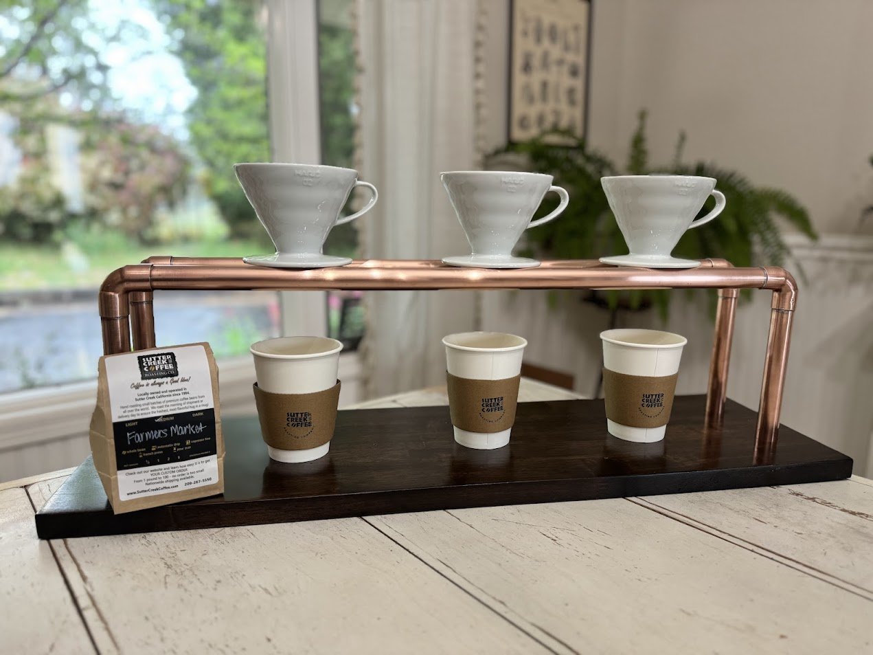 Coffee brewing setup with three white pour-over coffee drippers on a copper stand and three disposable coffee cups with cardboard sleeves on a dark wooden tray inside a house, with a window and greenery outside in the background.