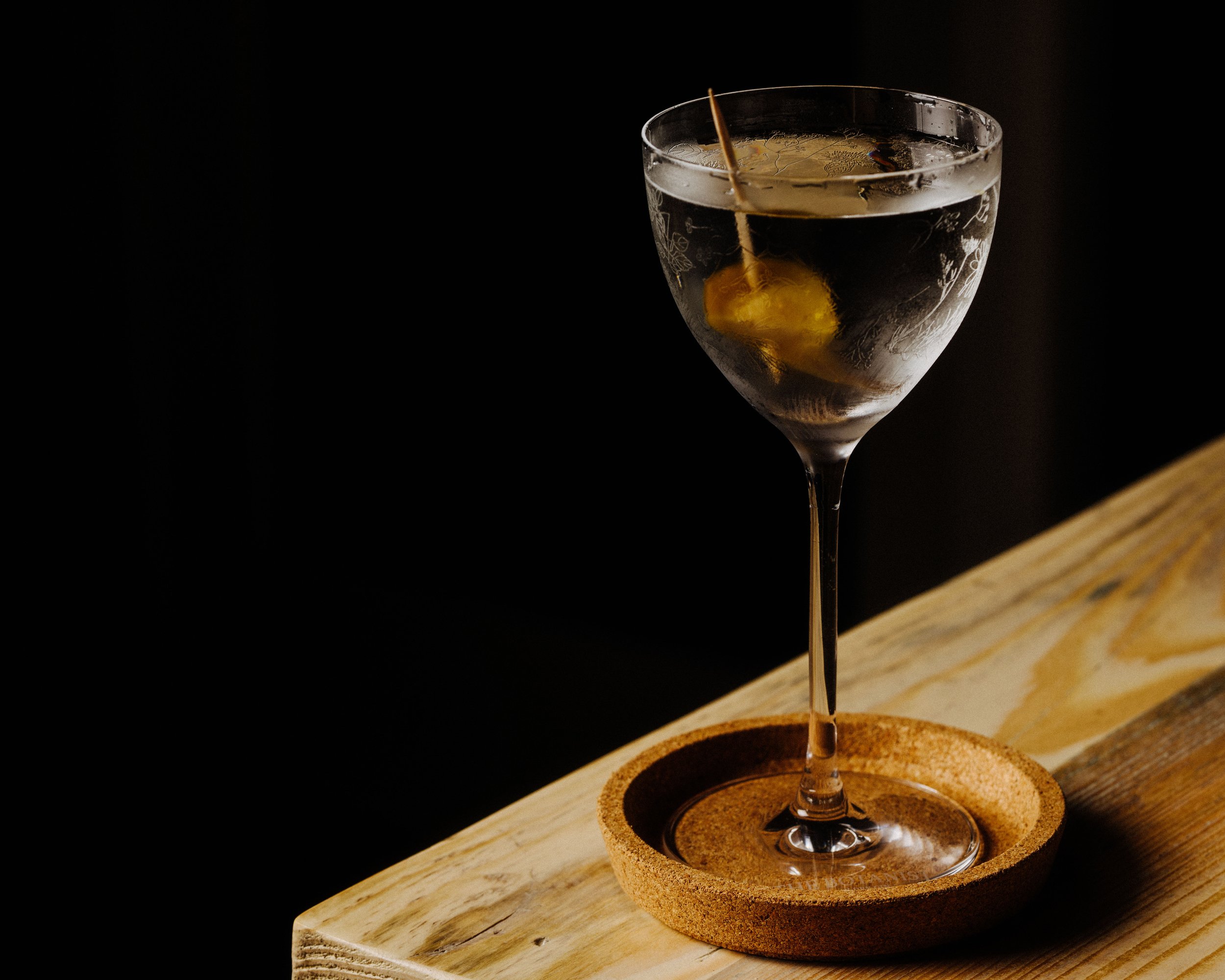 Fallachan Dining - A martini glass with a cocktail garnished with a lemon twist, placed on a cork coaster on a wooden table.
