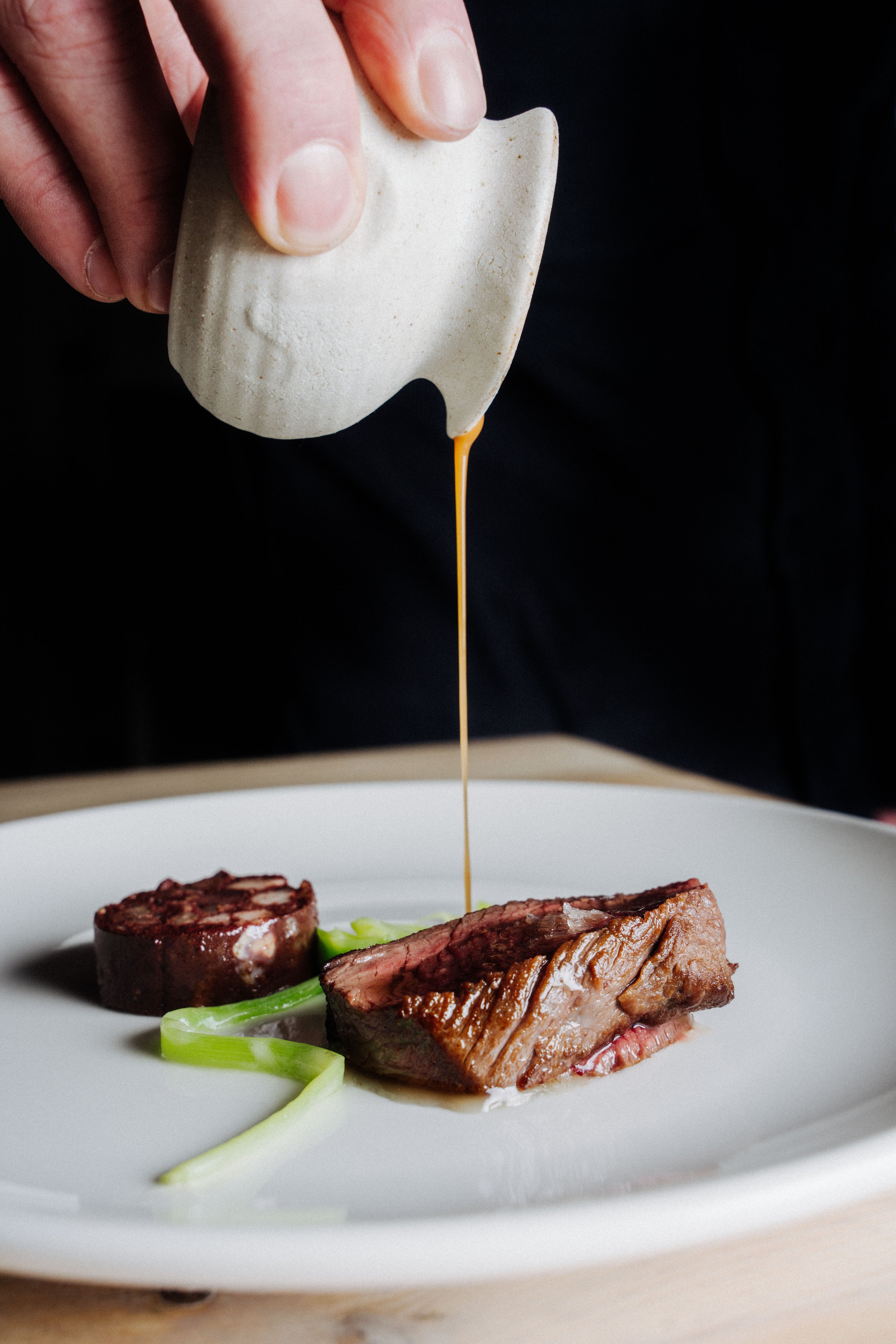 Fallachan Dining - A hand pouring sauce over a plate with cooked meat and vegetables.