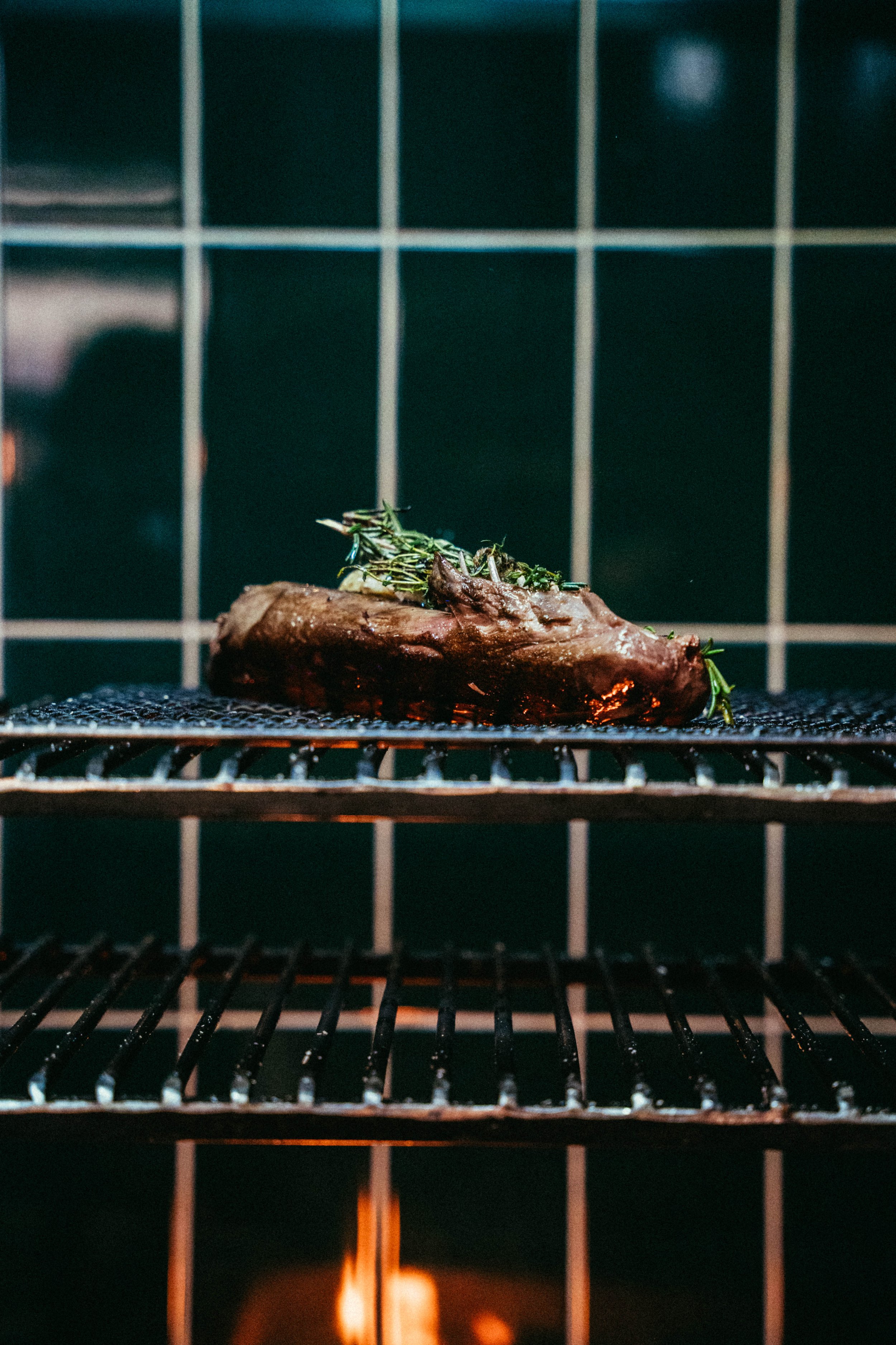 Fallachan Dining - Grilled steak with herbs on a barbecue grill with a flame below, tiled wall background.
