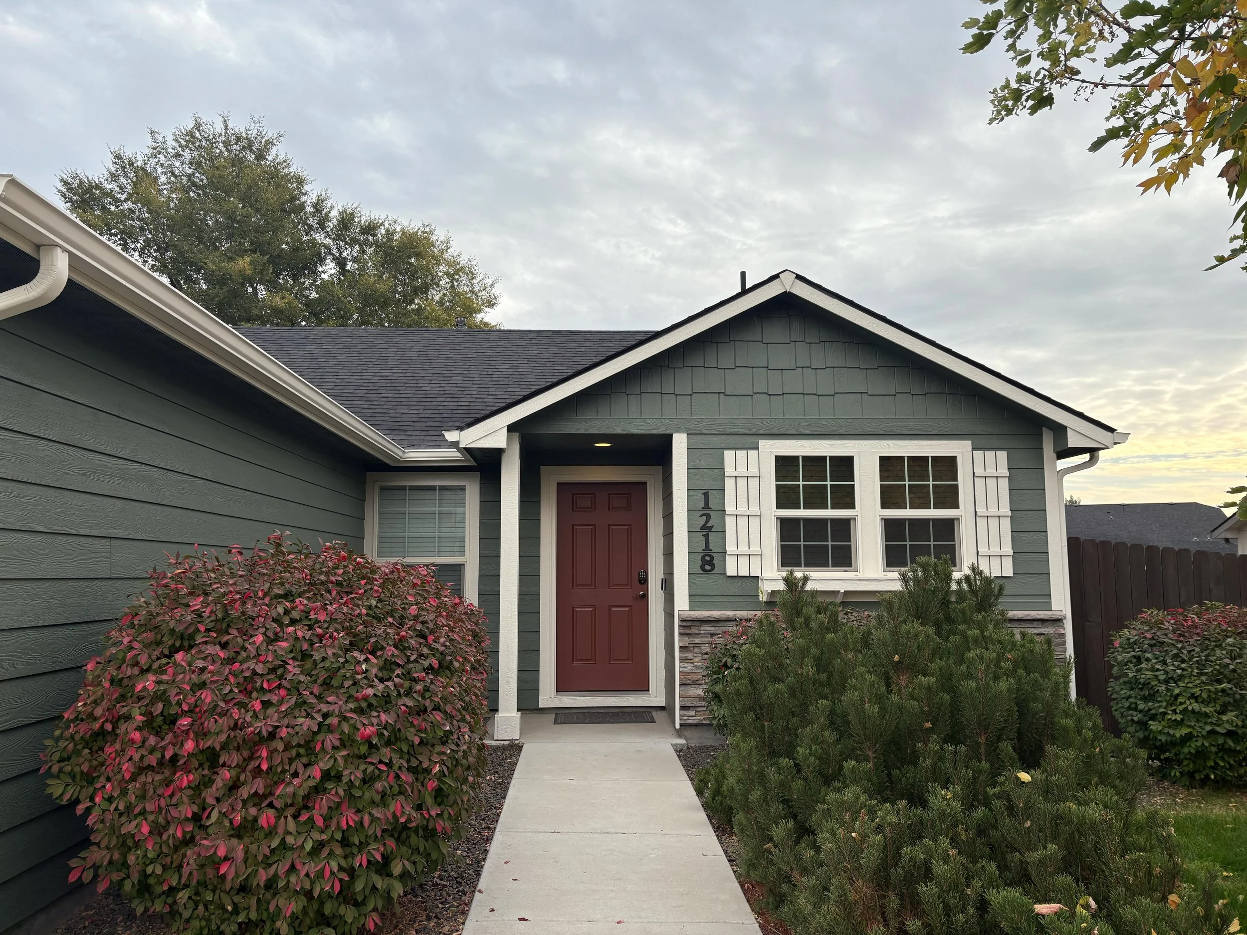 Front view of a single-story house with green siding, a brown front door, and white trim. There are bushes and shrubs in the landscaped front yard.
