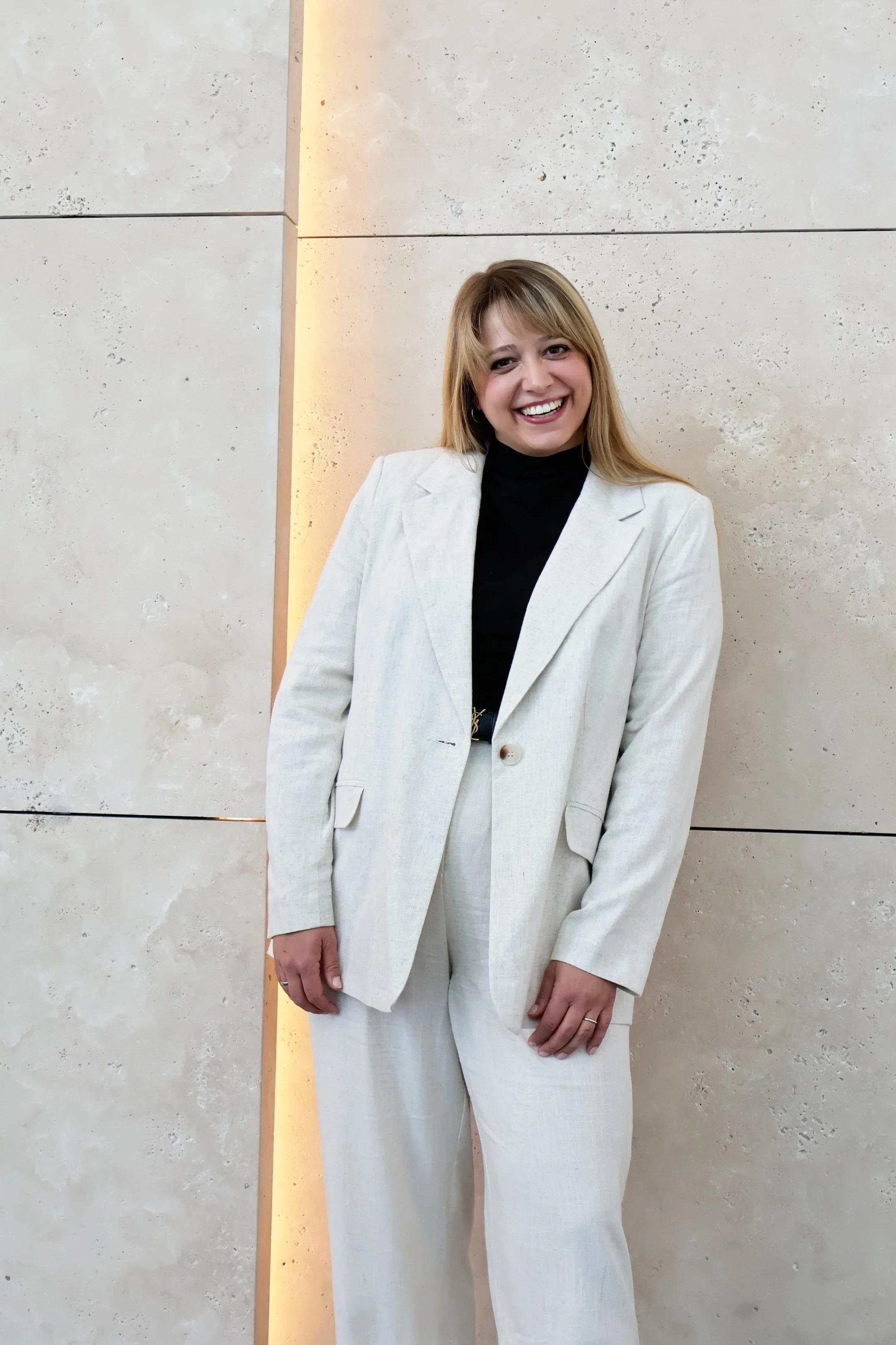 Junge Frau in weißem Anzug lächelnd vor einer hellen Steinwand.
