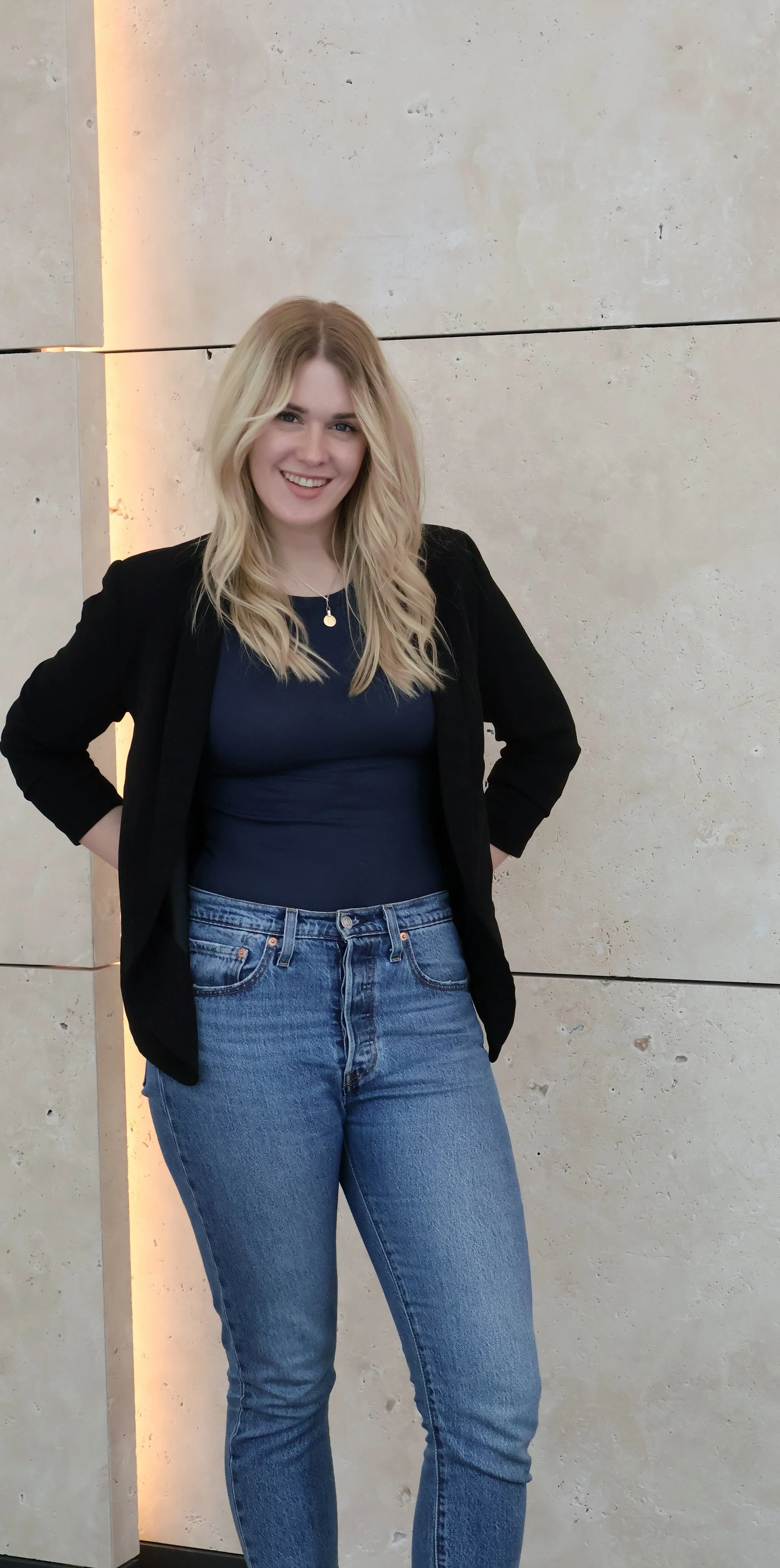 Junge Frau mit blonden, lockigen Haaren, schwarzer Jacke, dunklem Top und blauen Jeans, lächelnd vor einer hellen Steinwand.