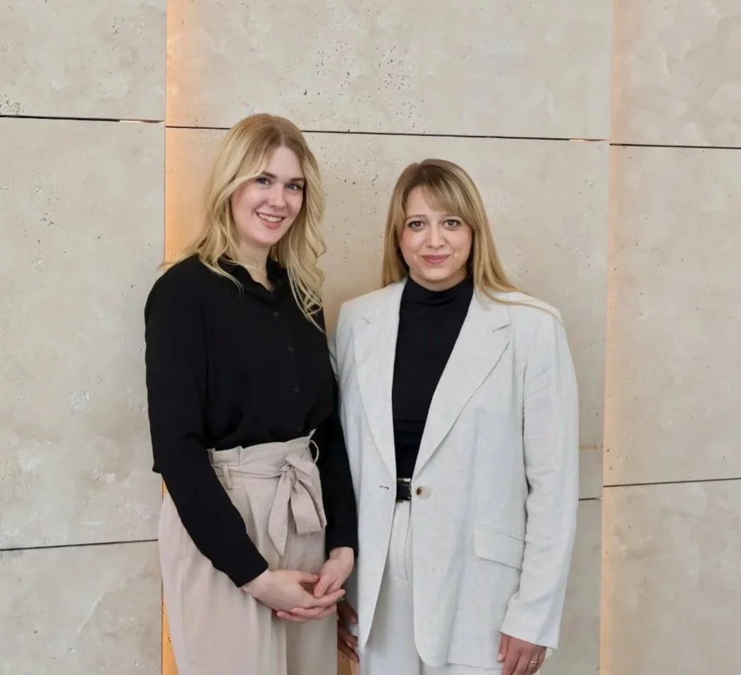 Zwei Frauen stehen vor einer hellen Steinwand, lächeln in die Kamera und posieren für ein Foto.