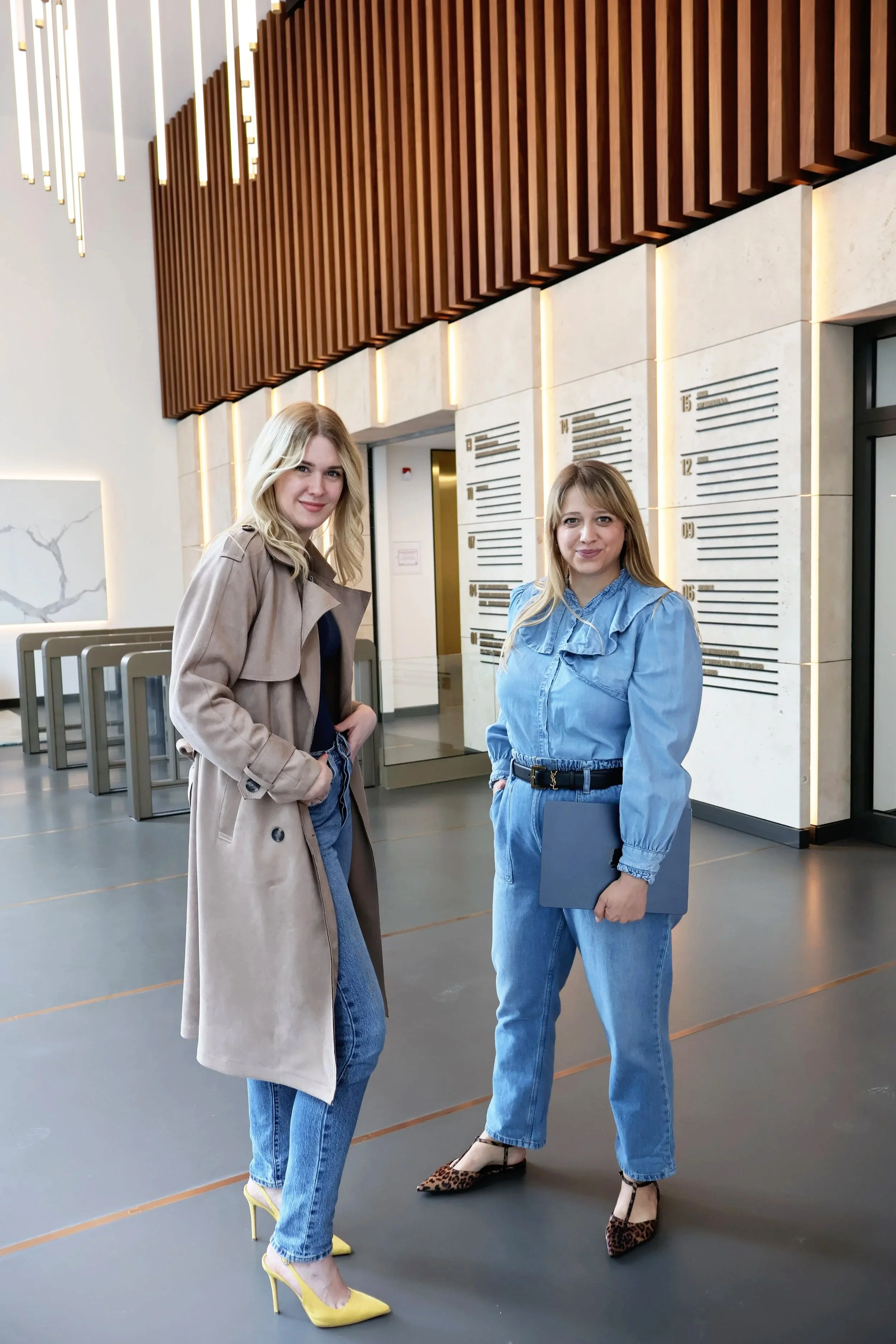 Zwei Frauen stehen in einem modernen Innenraum, eine trägt eine beige Trenchcoat und gelbe High Heels, die andere trägt einen blauen Overall und Leoparden-Print-High-Heels, beide lächeln und schauen in die Kamera.