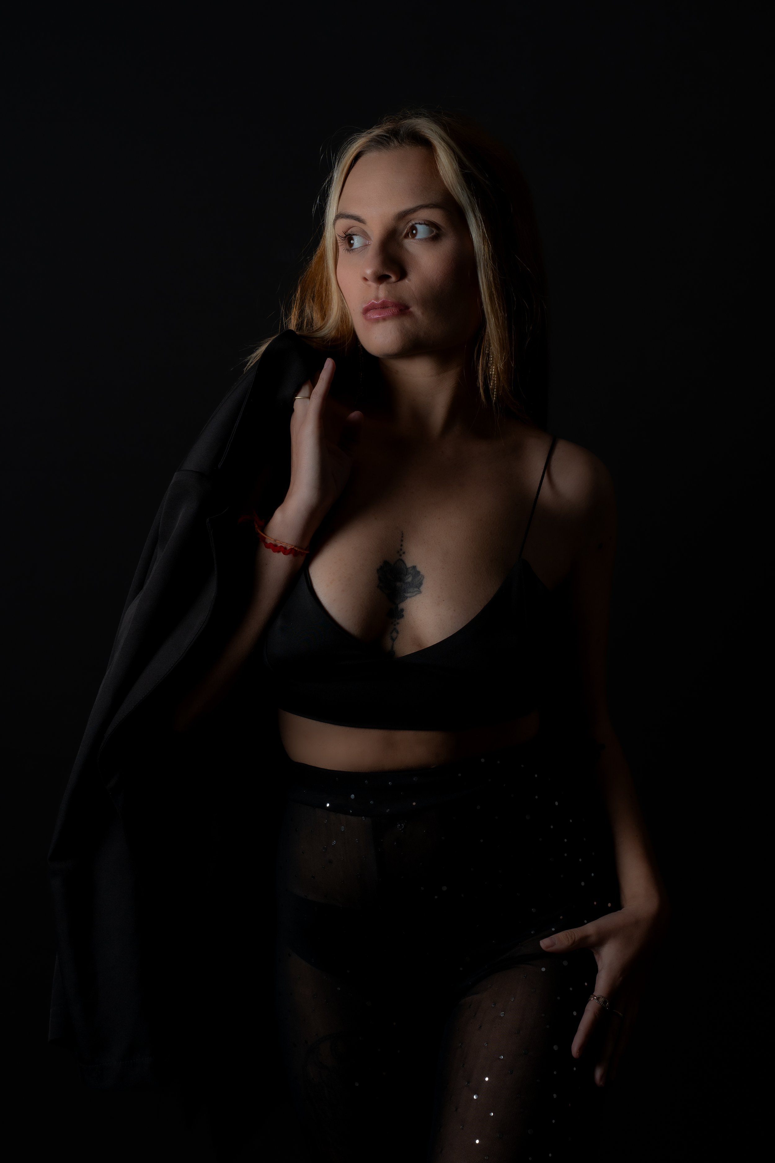 Femme posant dans un studio sombre, vêtue d'un haut noir et portant une veste sur l'épaule, avec un tatouage visible sur la poitrine.