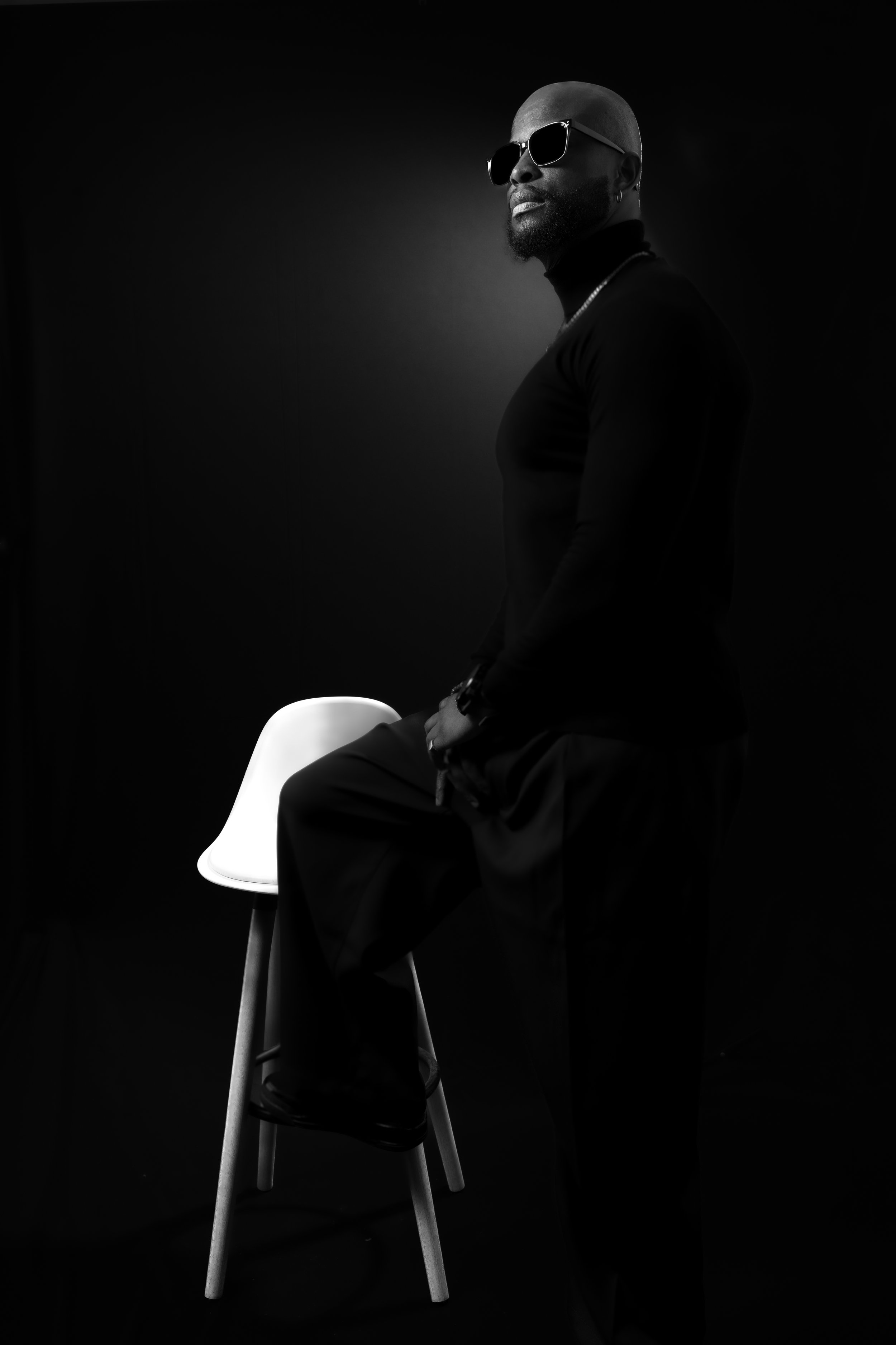 Homme avec lunettes de soleil, posant près d'une chaise blanche, fond noir.