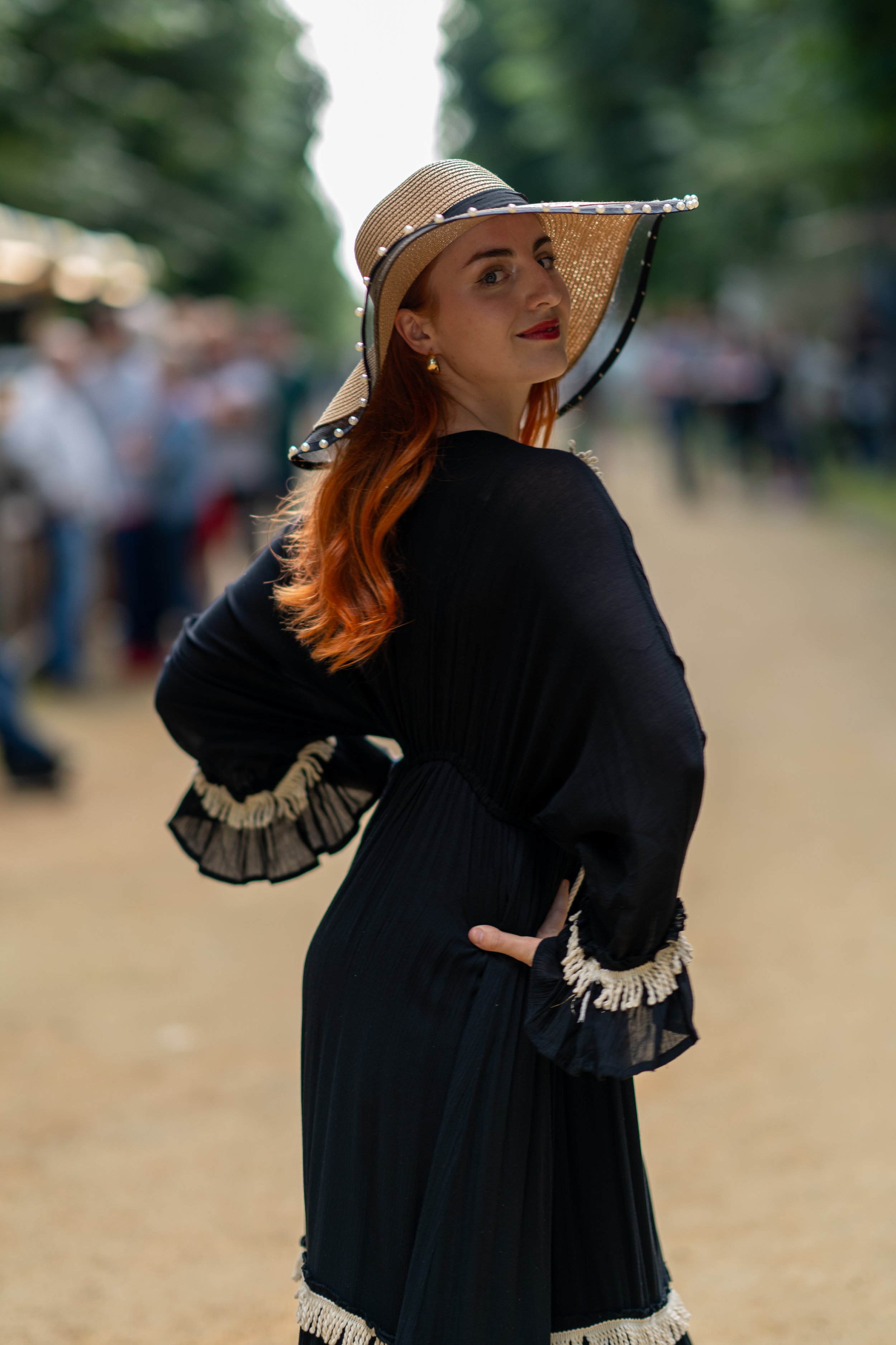 Femme portant une robe noire avec un chapeau élégant, posant à l'extérieur.