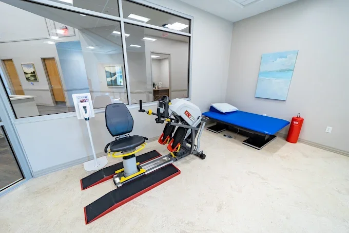 A medical rehabilitation room with an exercise machine, a bed with a pillow, and a fire extinguisher, with glass windows showing an adjacent office area.