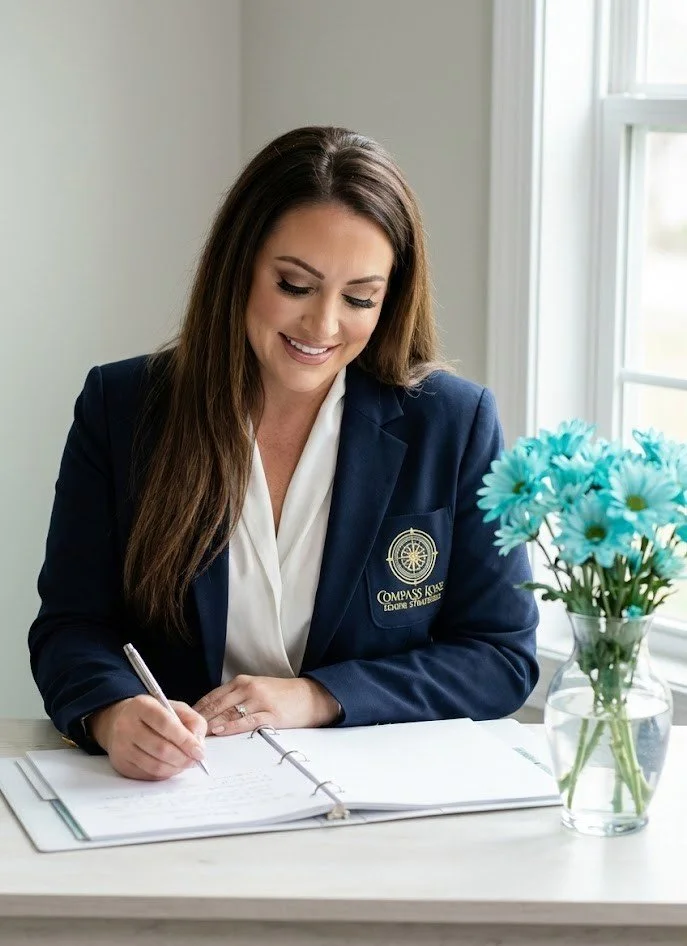 A woman in a navy blazer with a Compass logo writing in a binder at a desk near a window, with a vase of blue flowers.