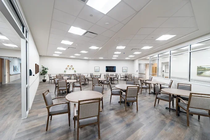 An empty, modern conference or waiting room with several round and rectangular tables, chairs, large windows, and wall-mounted screens.