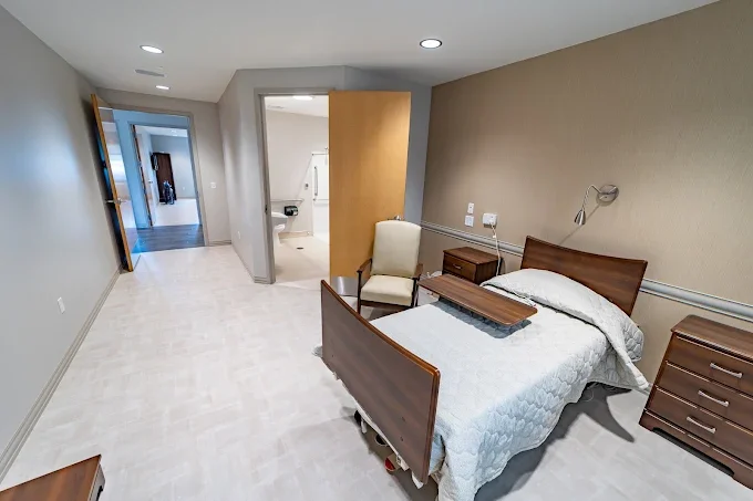 A hospital or nursing home room with a bed, a nightstand, an armchair, and a bathroom in the background.