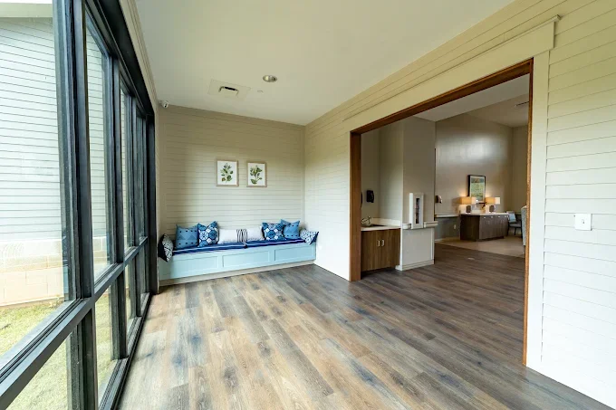 Empty sunroom with large windows, a built-in bench with blue pillows, framed botanical prints on the wall, and hardwood floors.