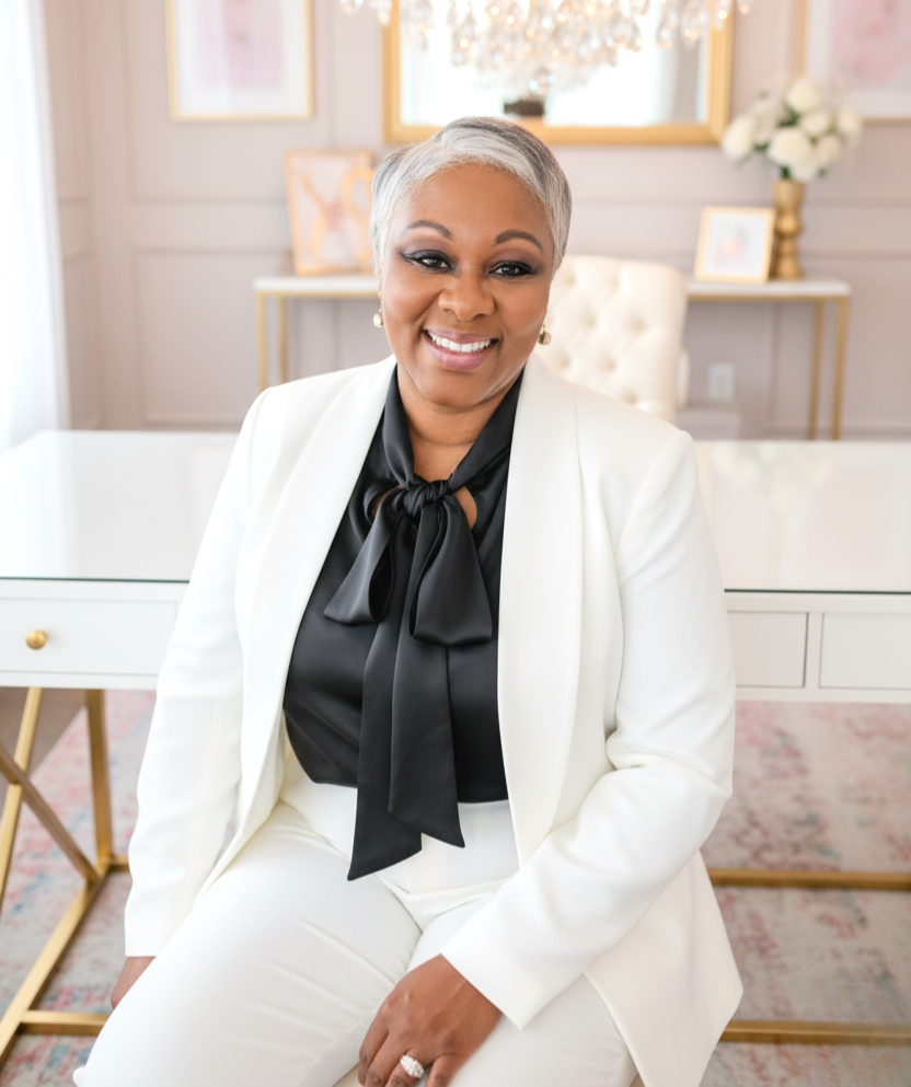A smiling woman with short gray hair wearing a white suit and black blouse with a bow, sitting in a bright, elegant office or living space with framed artwork, a chandelier, gold accents, and flowers in the background.