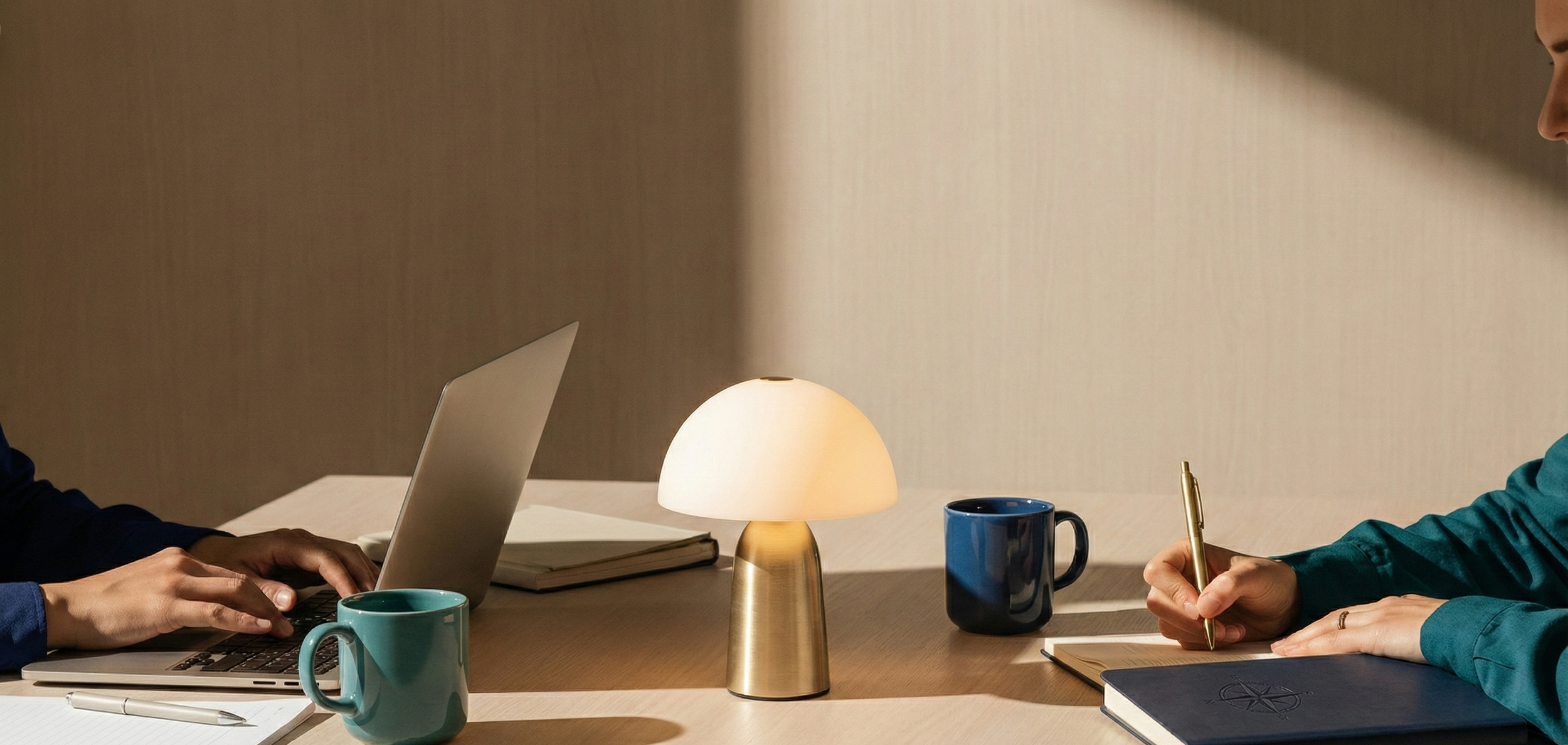 Two people working at a desk with a lamp, notebooks, a laptop, and mugs, one person is typing on a laptop, and the other is writing in a notebook.