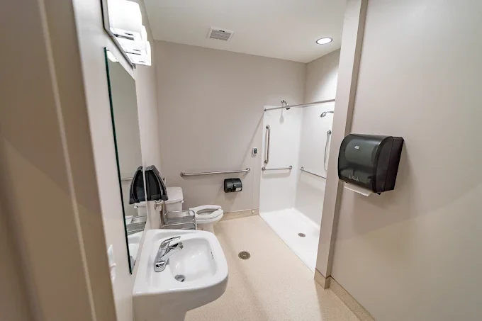 Accessible bathroom with sink, toilet, shower with grab bars, and a wall-mounted paper towel dispenser.