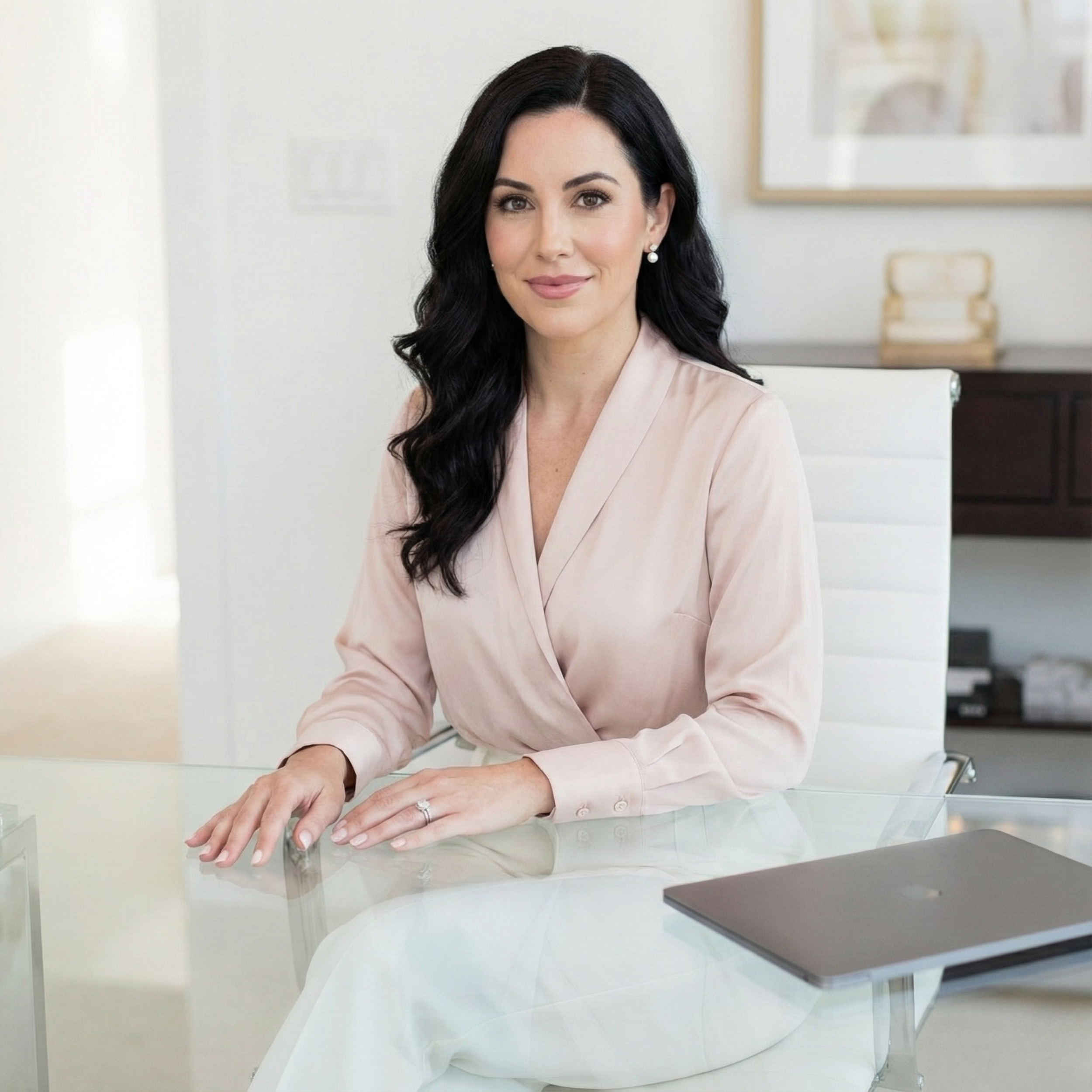 A woman with long dark hair, wearing a light pink blouse, sitting at a glass desk in a modern office, and looking at the camera.