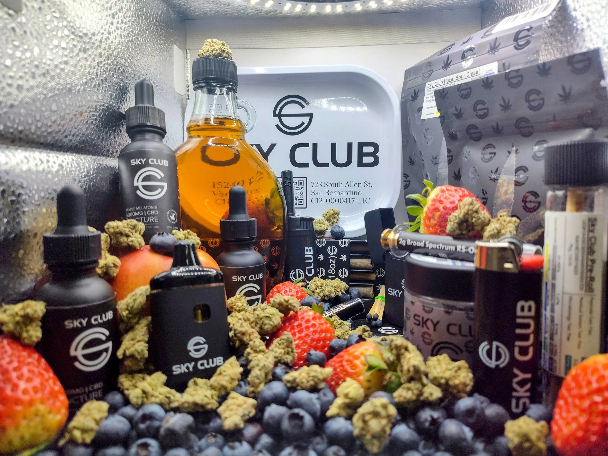 Inside a gray box, various cannabis products including bottles, jars, and bags with the Sky Club logo, surrounded by strawberries, blueberries, and cannabis buds.