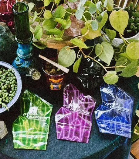 Three holographic packs labeled 'Crystals Collection' in green, pink, and blue are placed on a table surrounded by potted plants and other decorative items.