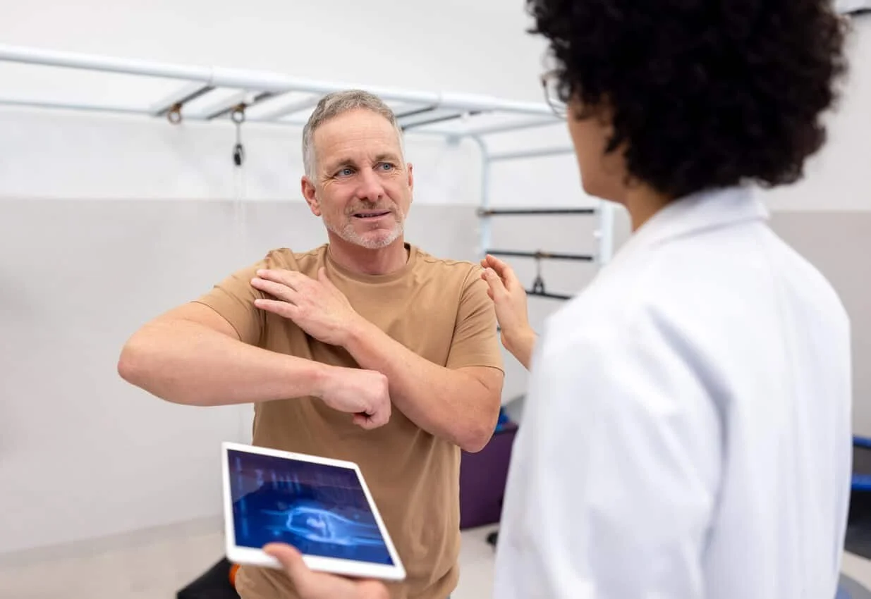 A middle-aged man in a beige t-shirt stretching his shoulder while talking to a woman in a white medical coat. The woman is holding a tablet displaying a medical image, and they are in a medical or physical therapy setting with exercise equipment in the background.