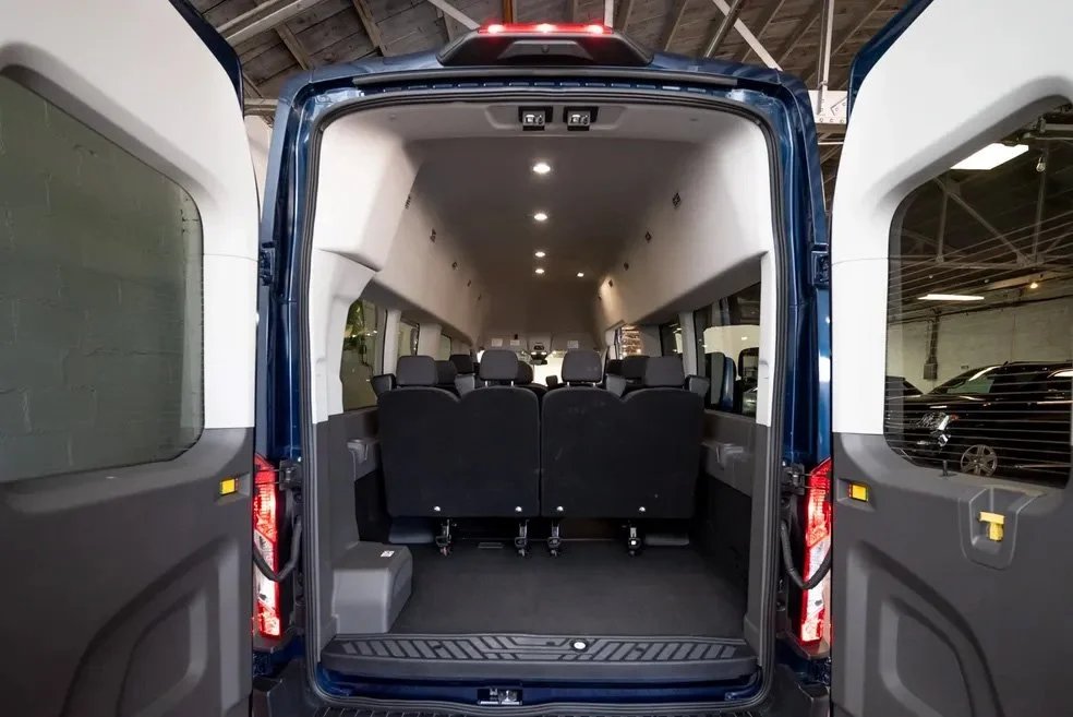 The interior of a passenger van with multiple rows of black seats, seen from the rear with doors open, inside a warehouse or garage.