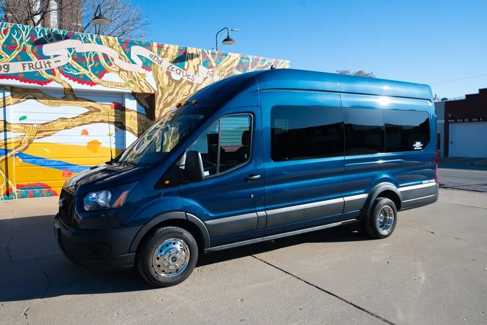 A dark blue passenger van parked on a street in front of a colorful mural with trees and buildings in the background.