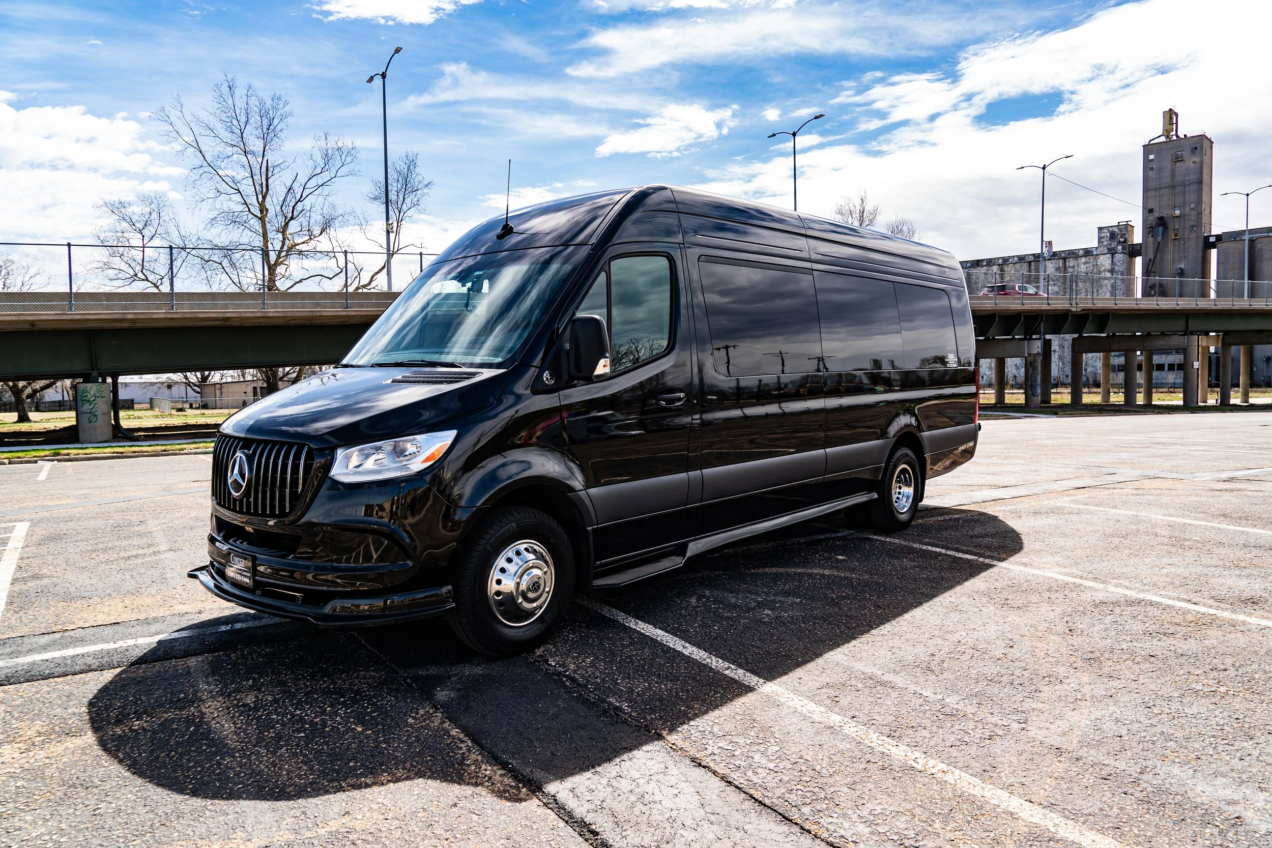Black Mercedes-Benz commercial van parked in an empty parking lot under a partly cloudy sky.