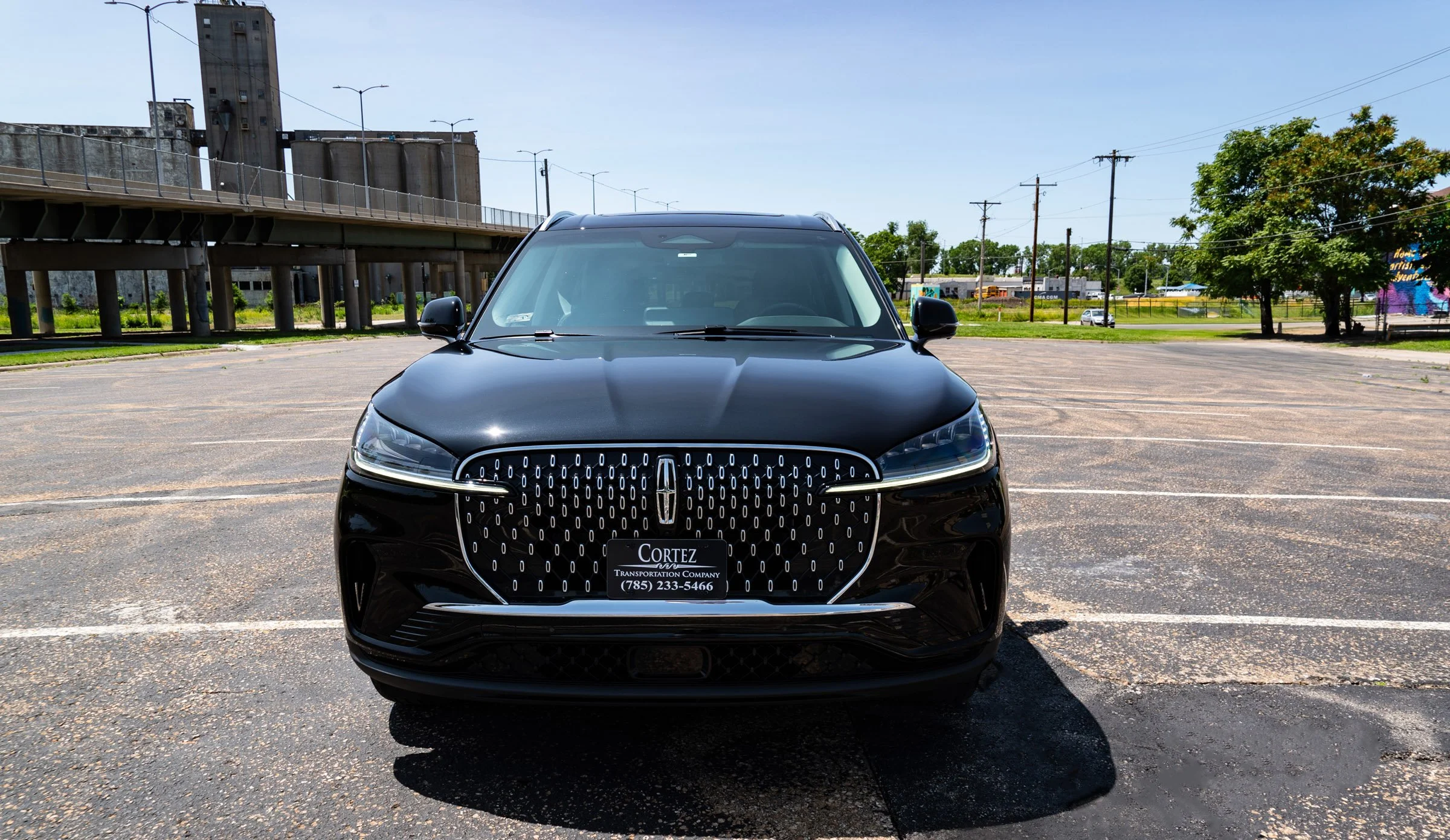 Black electric SUV parked in an empty lot with a custom front grille and corporate license plate, in an urban setting with trees, utility poles, and a bridge in the background.