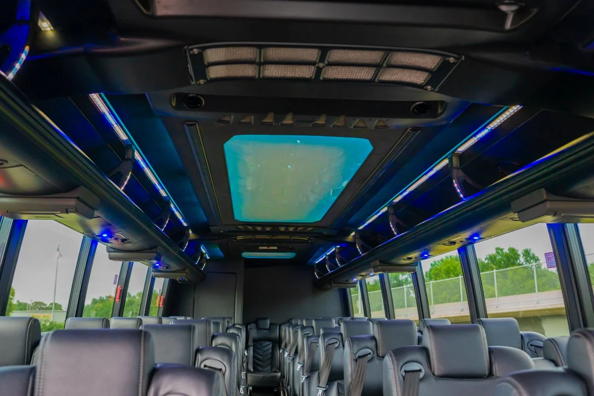 Interior of a luxury bus with leather seats, large windows, and a ceiling-mounted entertainment screen surrounded by blue LED lights.