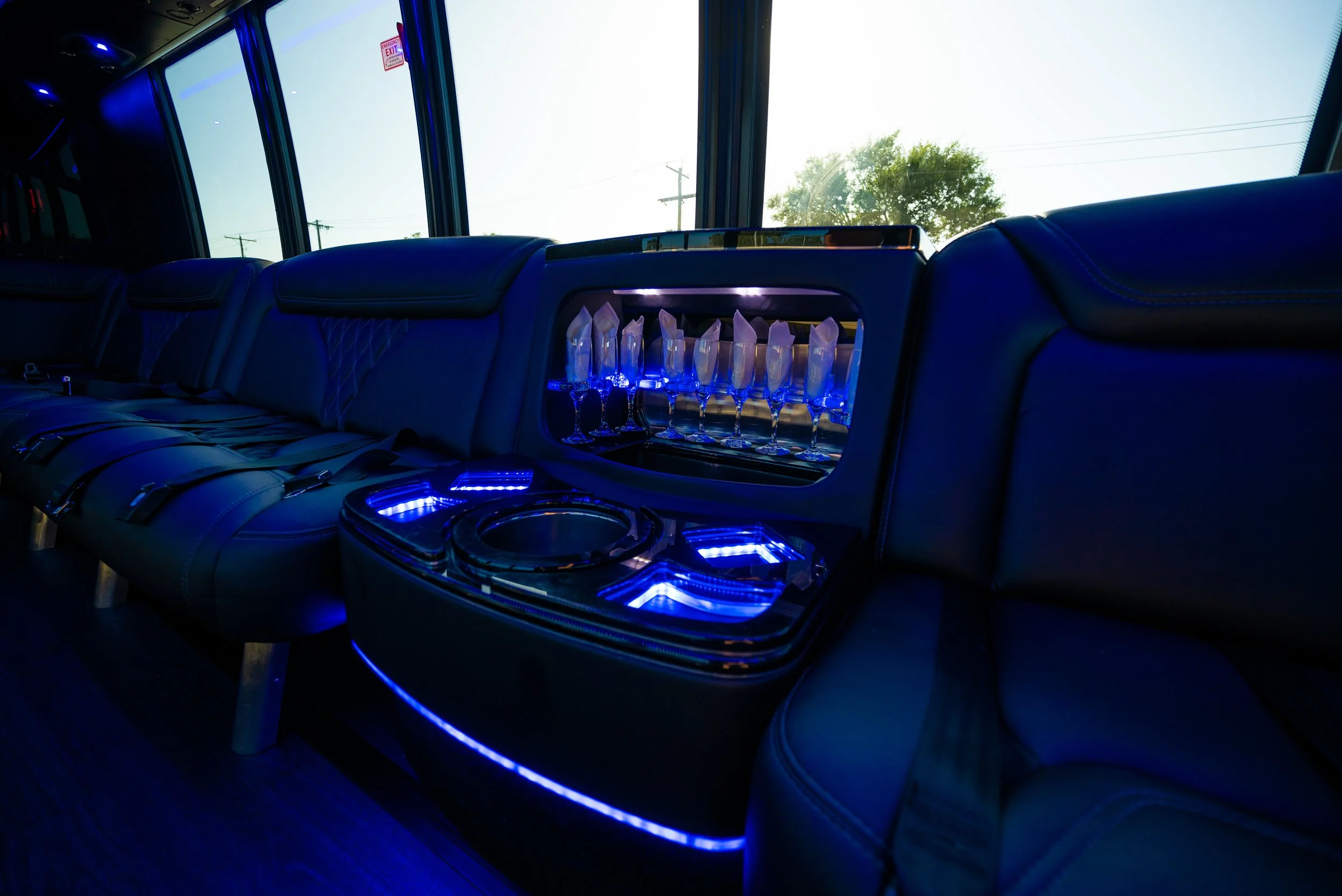 Luxury bus interior with black leather seats, a built-in bar with crystal champagne glasses, blue LED lighting, large windows showing trees and power lines outside.