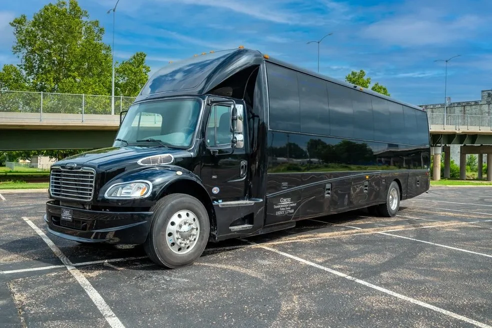 Black shuttle bus parked in a lot with a bridge and green trees in the background.