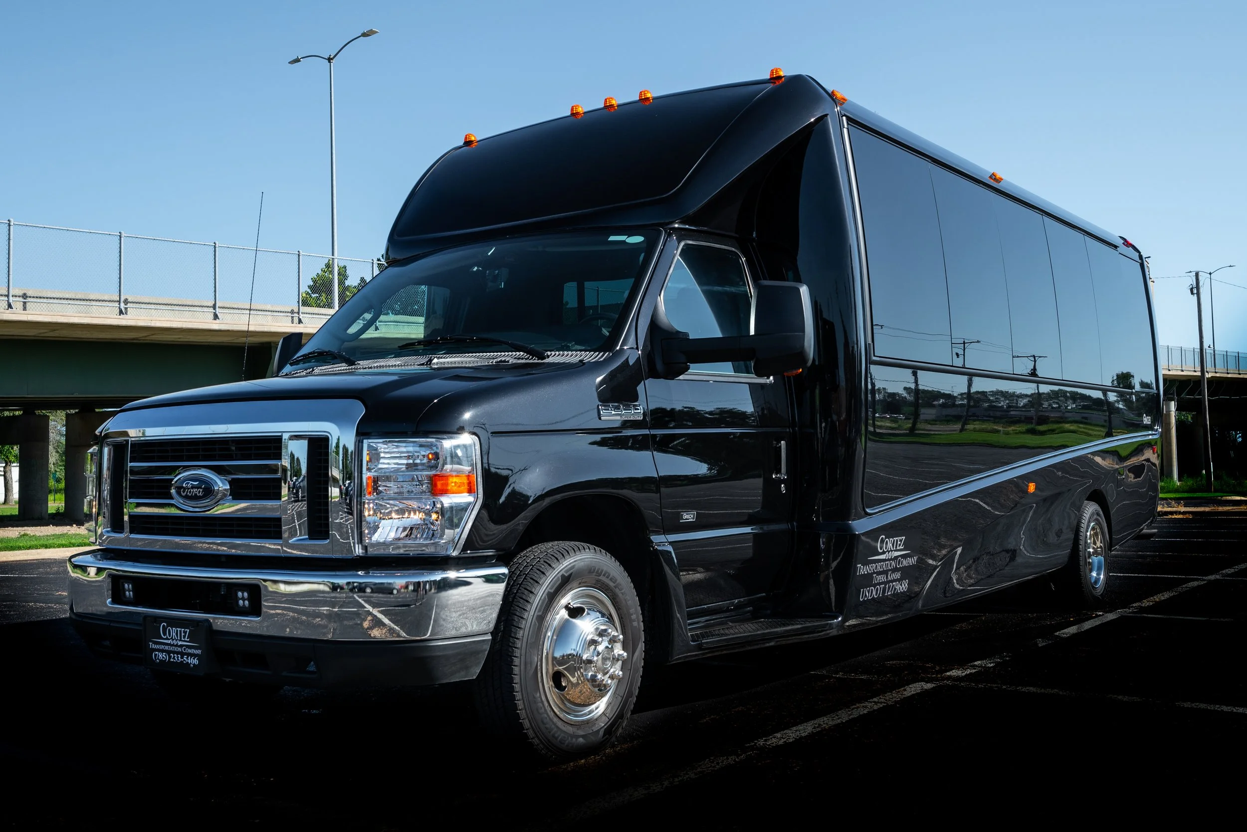 Black shuttle bus parked in a lot under a clear blue sky with a bridge in the background.