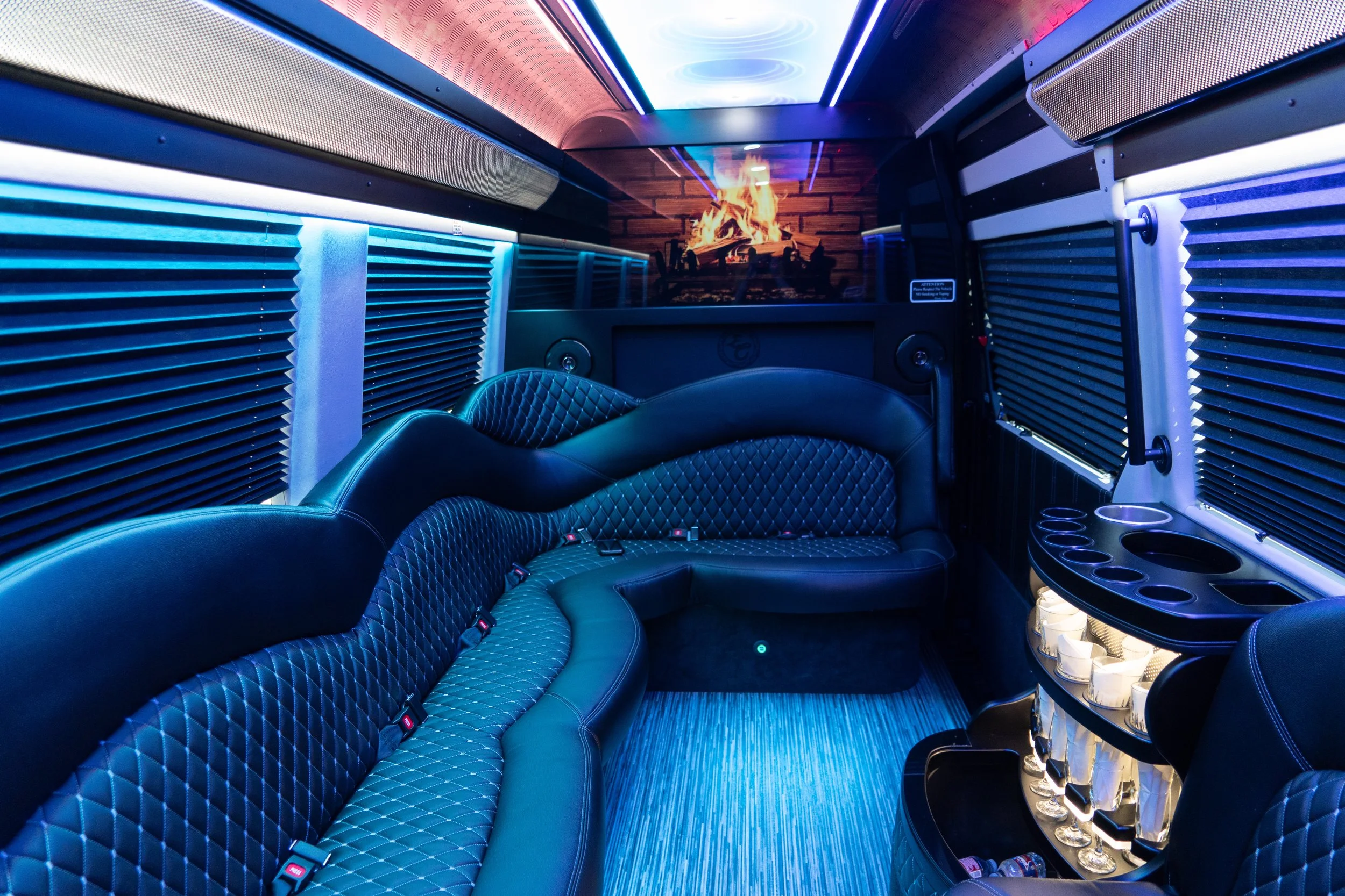 Luxury party bus interior with black quilted leather seating, blue lighting, a small bar area with glasses and beverages, and a fireplace mural.