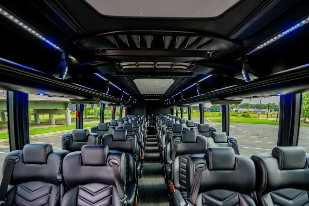 Interior of a bus with multiple rows of black leather seats and large windows, showing an outdoor parking lot and green trees.