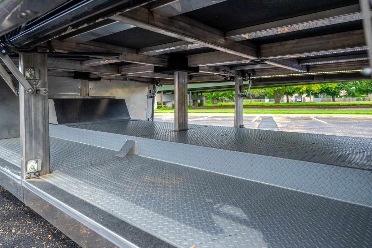 Underside of a metallic truck or trailer with a textured metal floor and support beams, parked in an outdoor lot with trees and grass in the background.