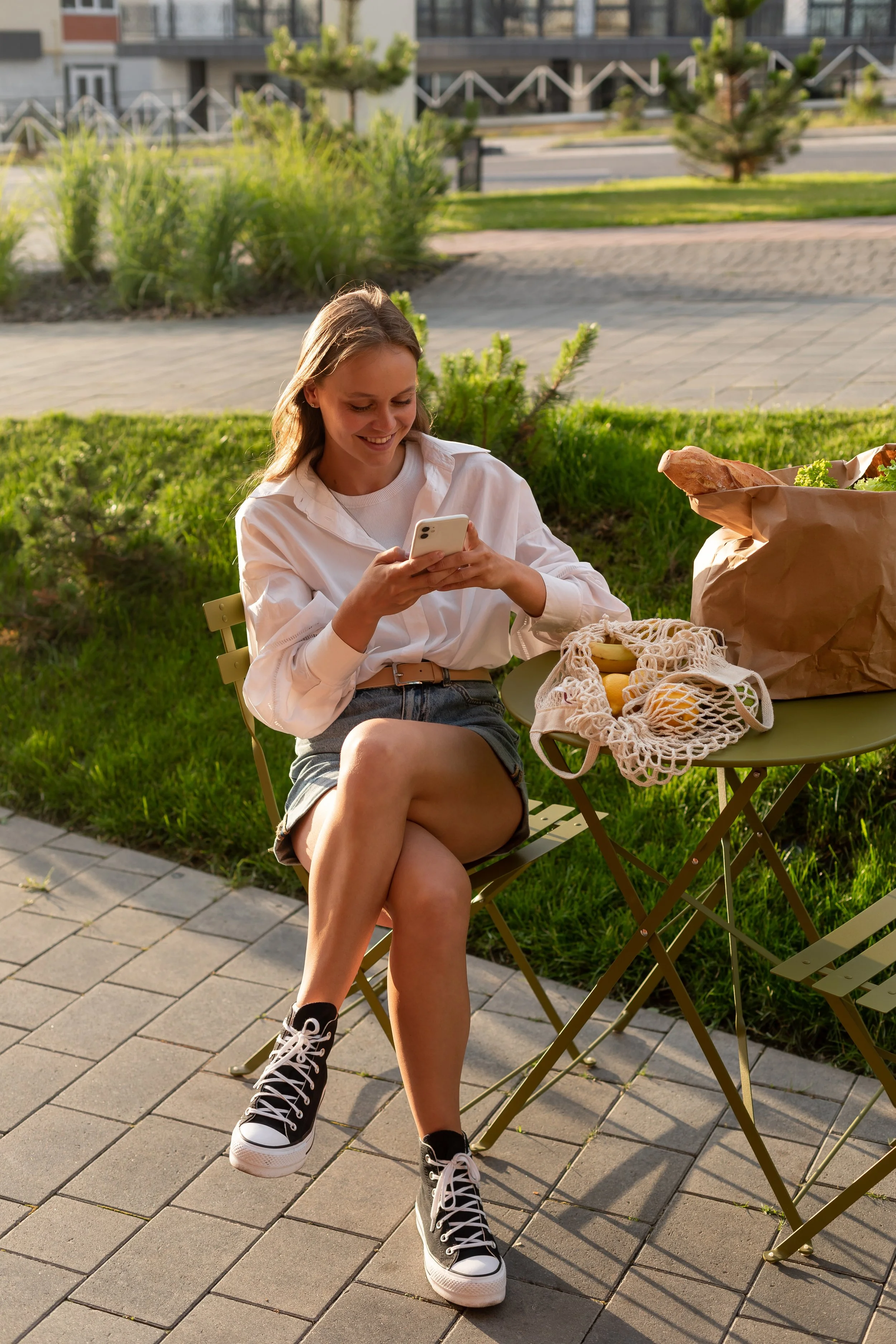 A young woman sitting outside on a folding chair, looking at her smartphone, with a basket of bananas and a paper bag on a green table next to her, surrounded by greenery.