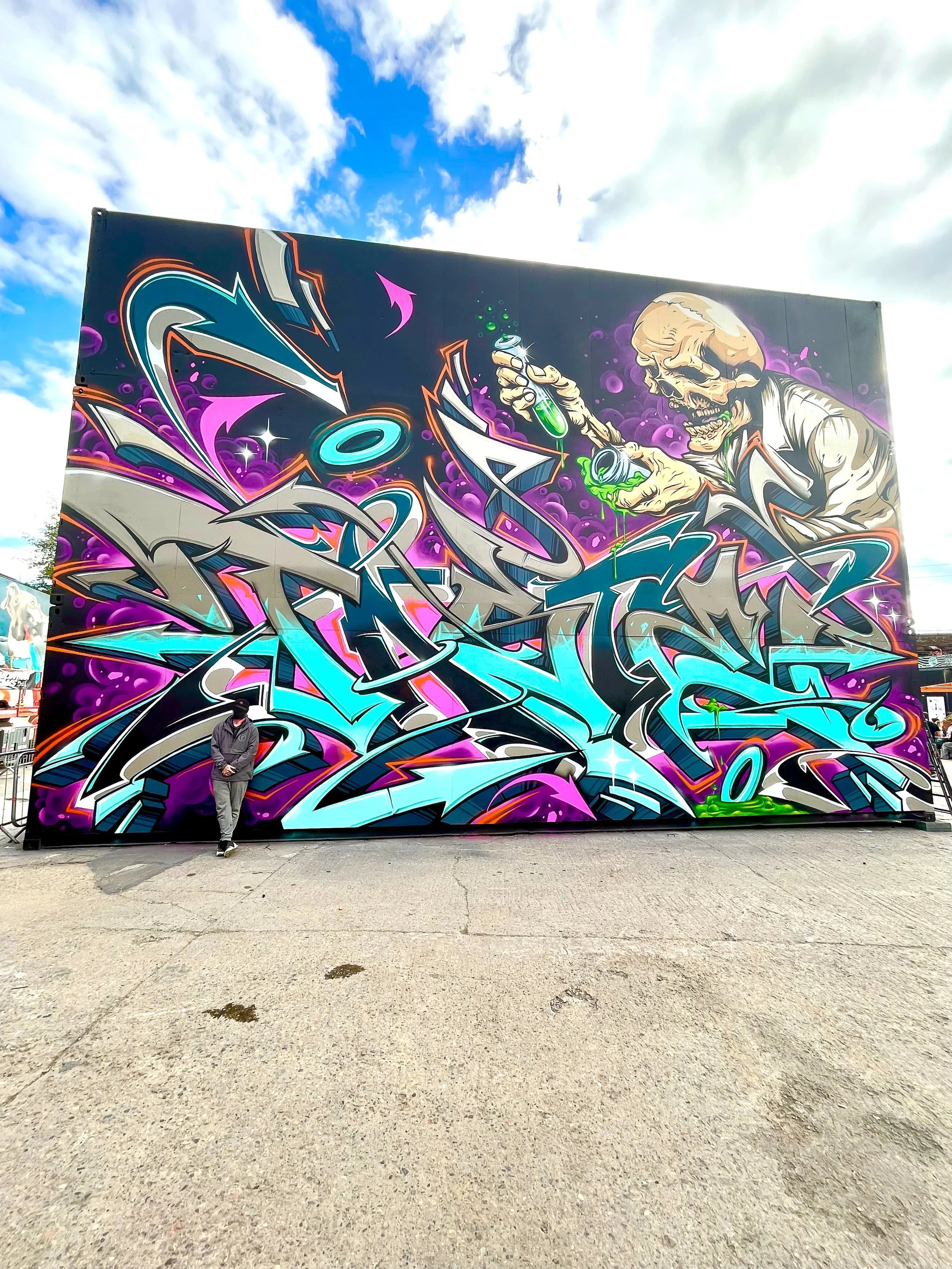 A photo of a graffiti artist standing in front of his massive wildstyle mural that depicts a large bright colored graffiti burner and an animated illustration of a mad scientist skeleton with a sky visible in the background