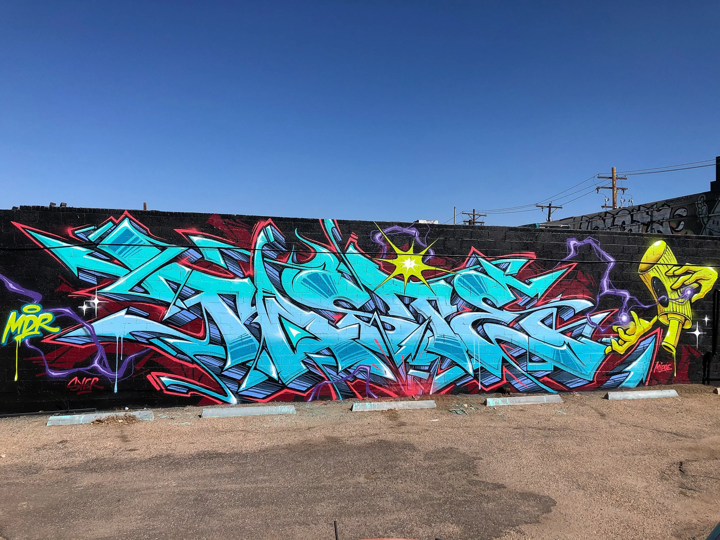 A large blue graffiti mural on a black brick depicting a spray cap character set against a clear blue sky in the background