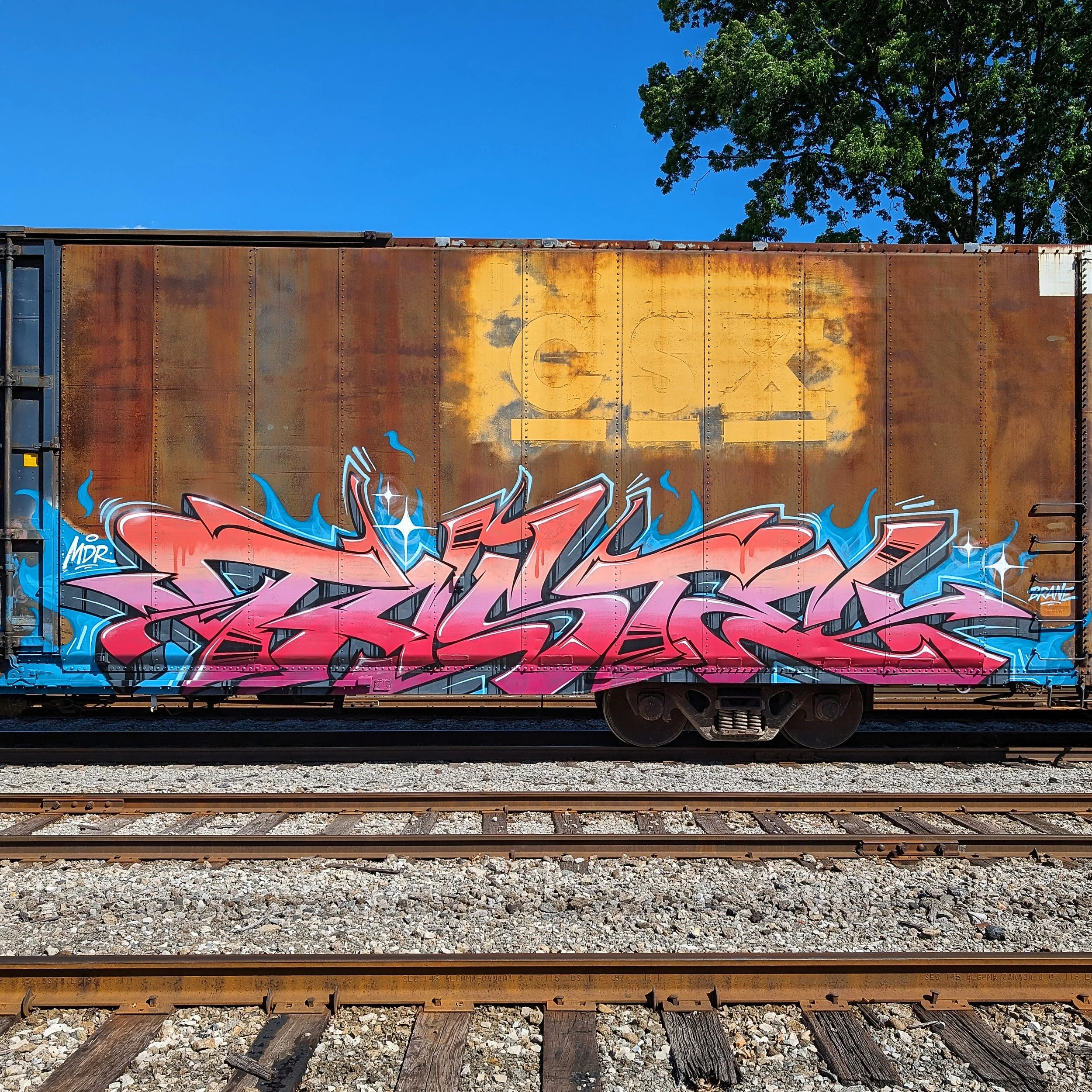 vibrant graffiti lettering on the side of a rusty CSX traincar witha  clear blue sky in the background of the train yard