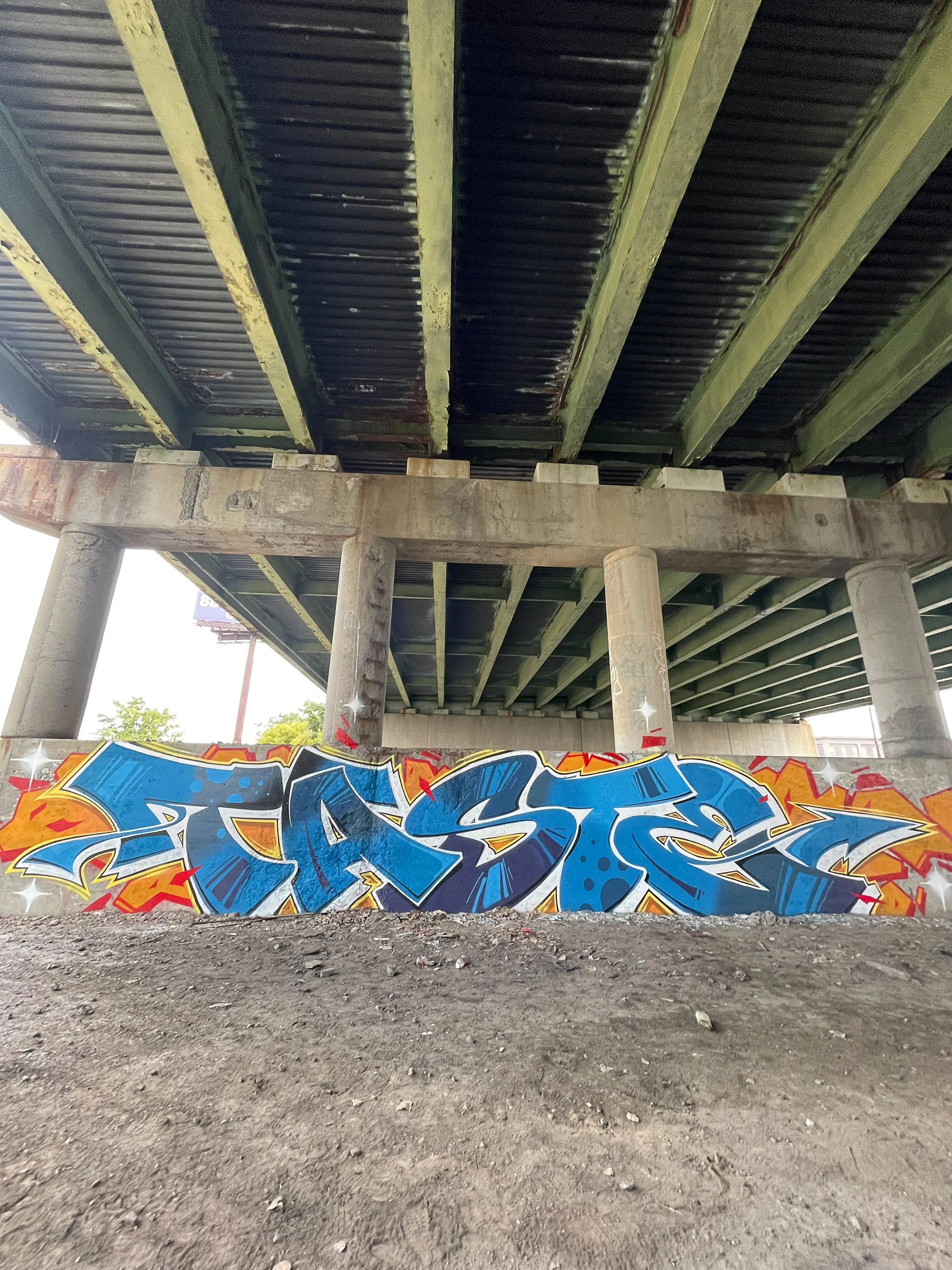 large graffiti lettering mural underneath bridge pillars on large walls surrounded by rocks and train tracks