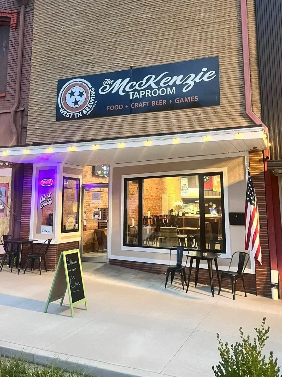 Exterior of a taproom and brewery named The MoKennzie Taproom, with outdoor seating, a chalkboard sign, and an American flag outside.