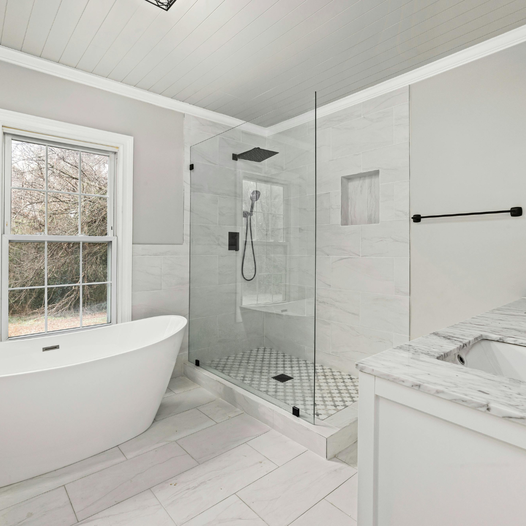 Modern bathroom with white bathtub, glass-enclosed shower, and marble countertop, featuring a window and neutral tile flooring.