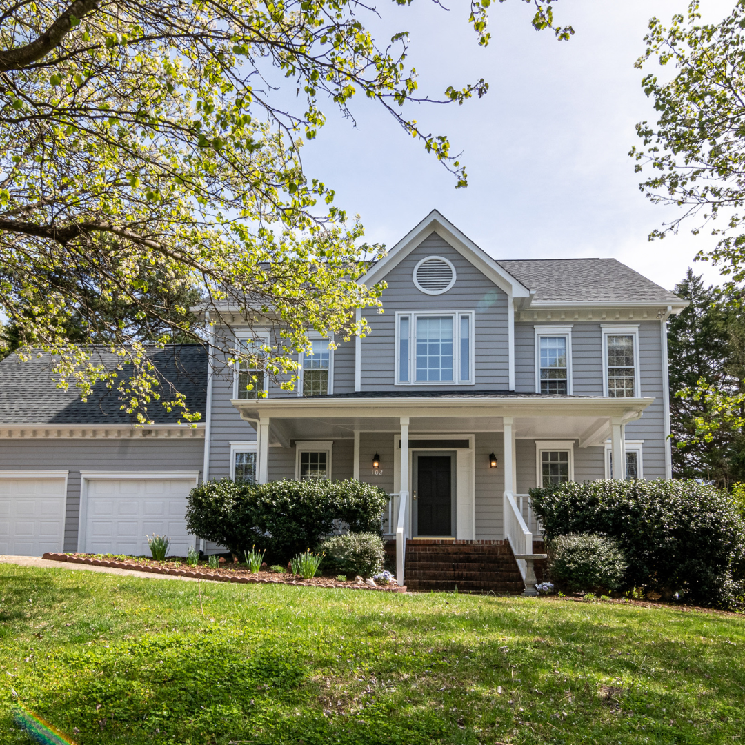 A two-story house with gray siding, a front porch with white columns, and steps leading to a black front door. The house has multiple windows and a gabled roof. There are bushes and small plants in the front yard, and a large tree with green leaves o