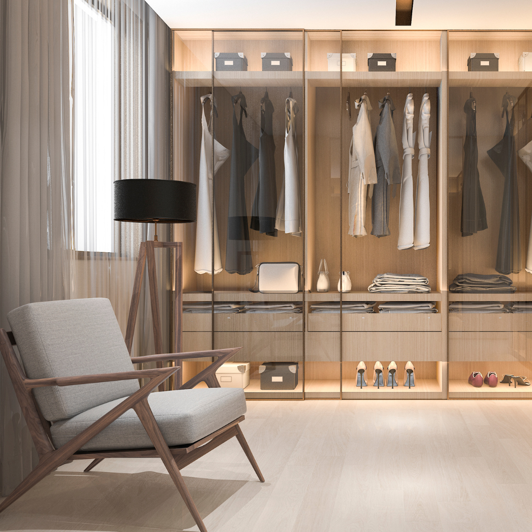 A modern walk-in closet with glass doors, containing hanging clothes, shelves with folded items, and shoes. There is a gray armchair and a black lamp on a wooden tripod nearby.