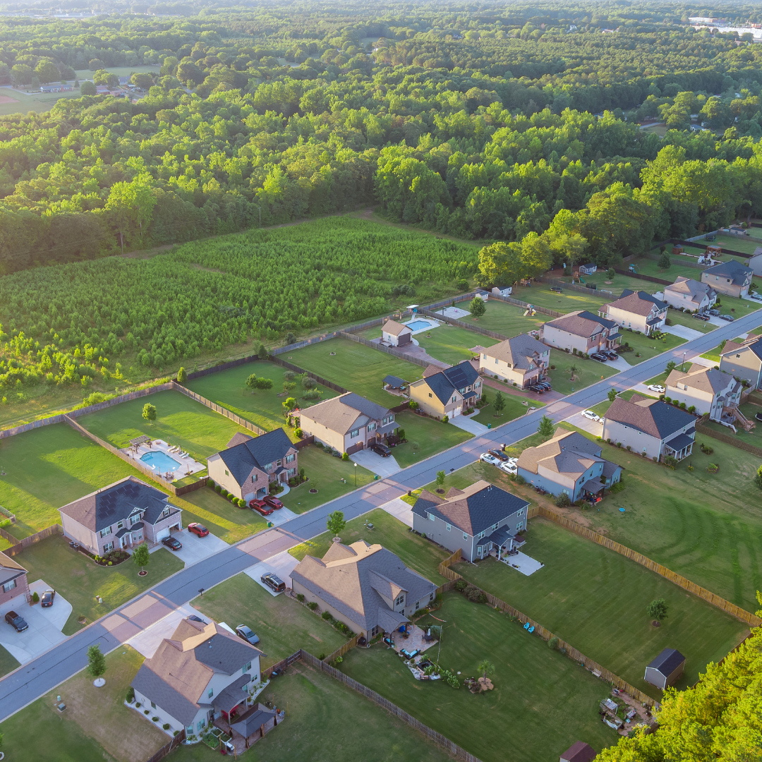 Aerial view of a suburban neighborhood with houses, lawns, and fenced backyards, some with swimming pools, bordered by a large green field and dense forest in the background.