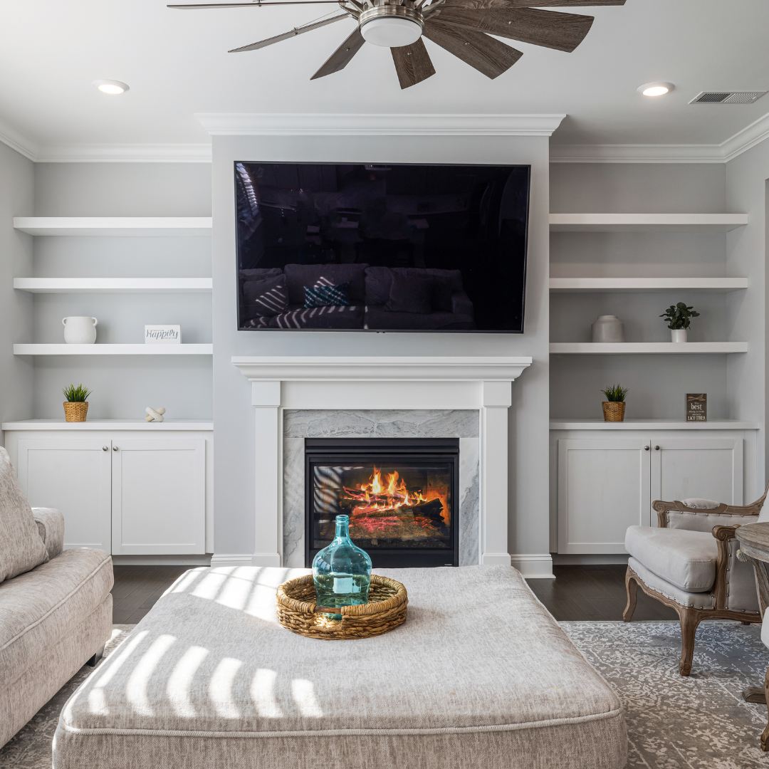 Living room with a fireplace, mounted TV above it, white built-in shelves on each side, beige sofa, and armchair, coffee table with a glass bottle in a woven tray, ceiling fan, and natural light