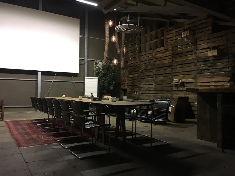 Een vergaderruimte met een lange tafel, zwarte stoelen, een whiteboard, een groot wit scherm, en decoratieve lampen, met een houten muur op de achtergrond.