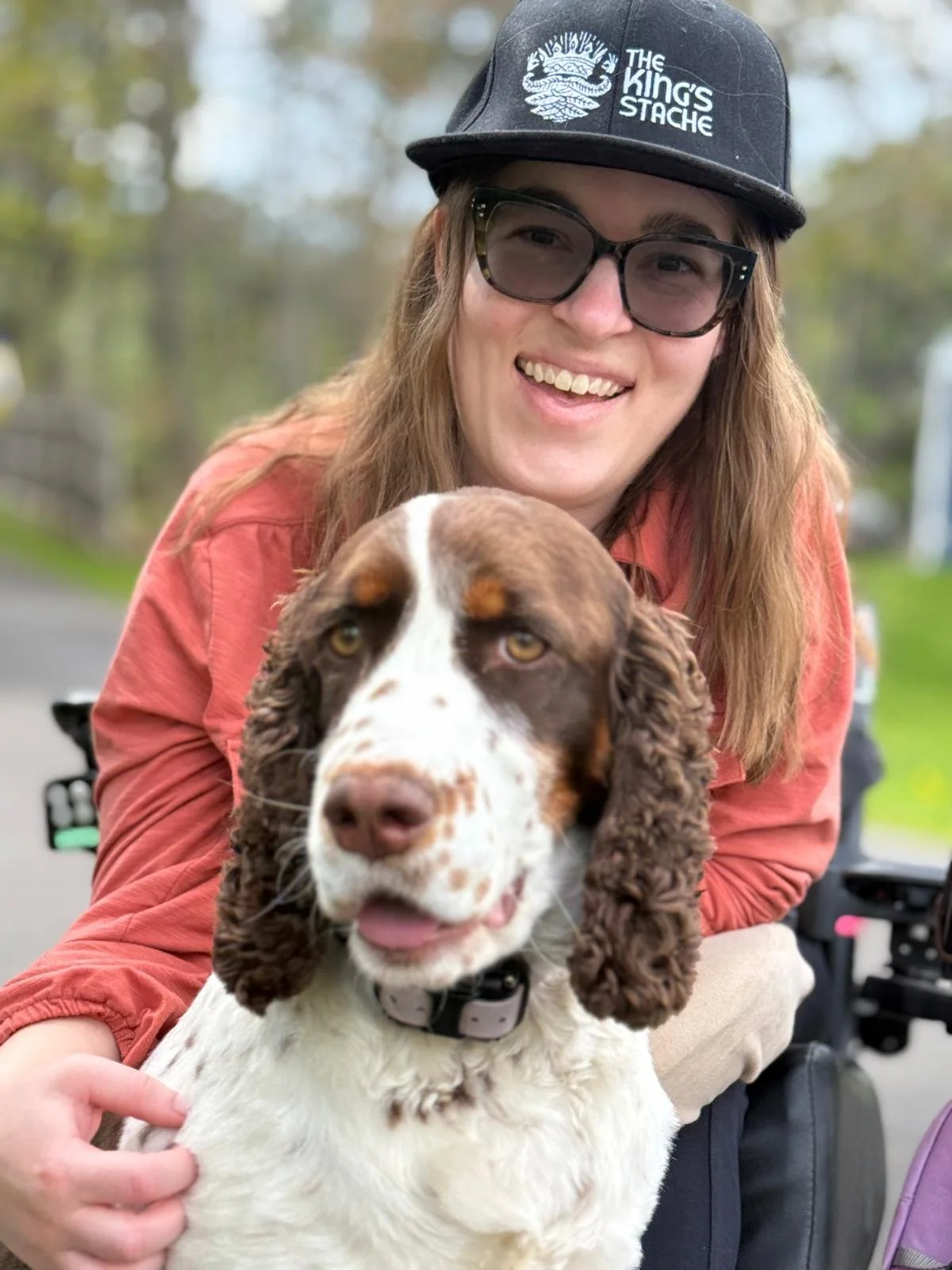 Image Description: Kings Presents as a white woman in a power chair, wearing a rust colored shirt and black hat. The hat says "The King's Stache" on it with a crown and leaves. In front of Kings is her dog, Zadie, a liver and white English Springer.
