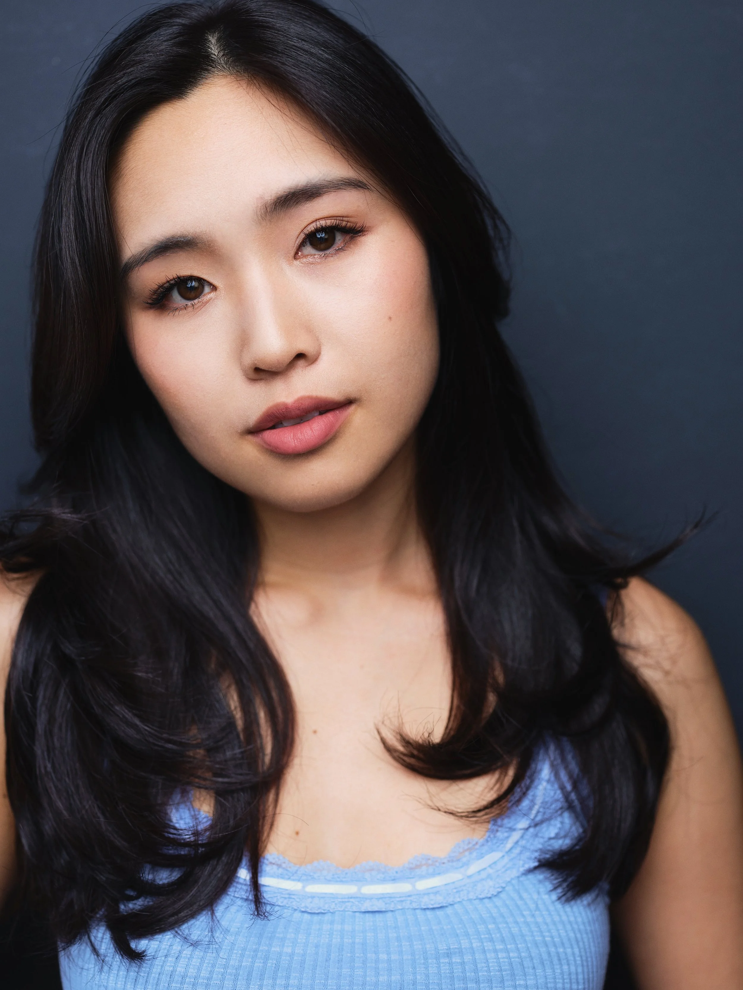 Portrait of a young woman with black hair, brown eyes, and light skin, wearing a blue top, against a dark background.
